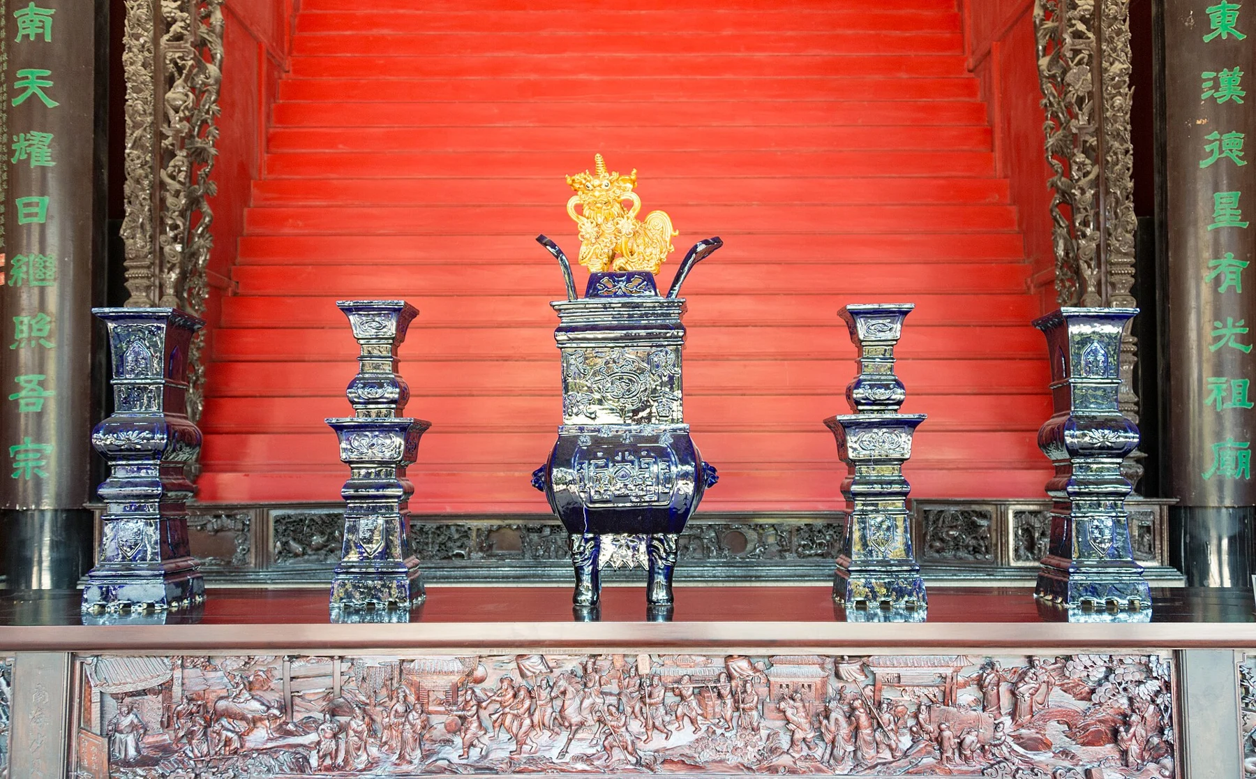Blue decorated vessels and central ornament before a red hall backdrop.