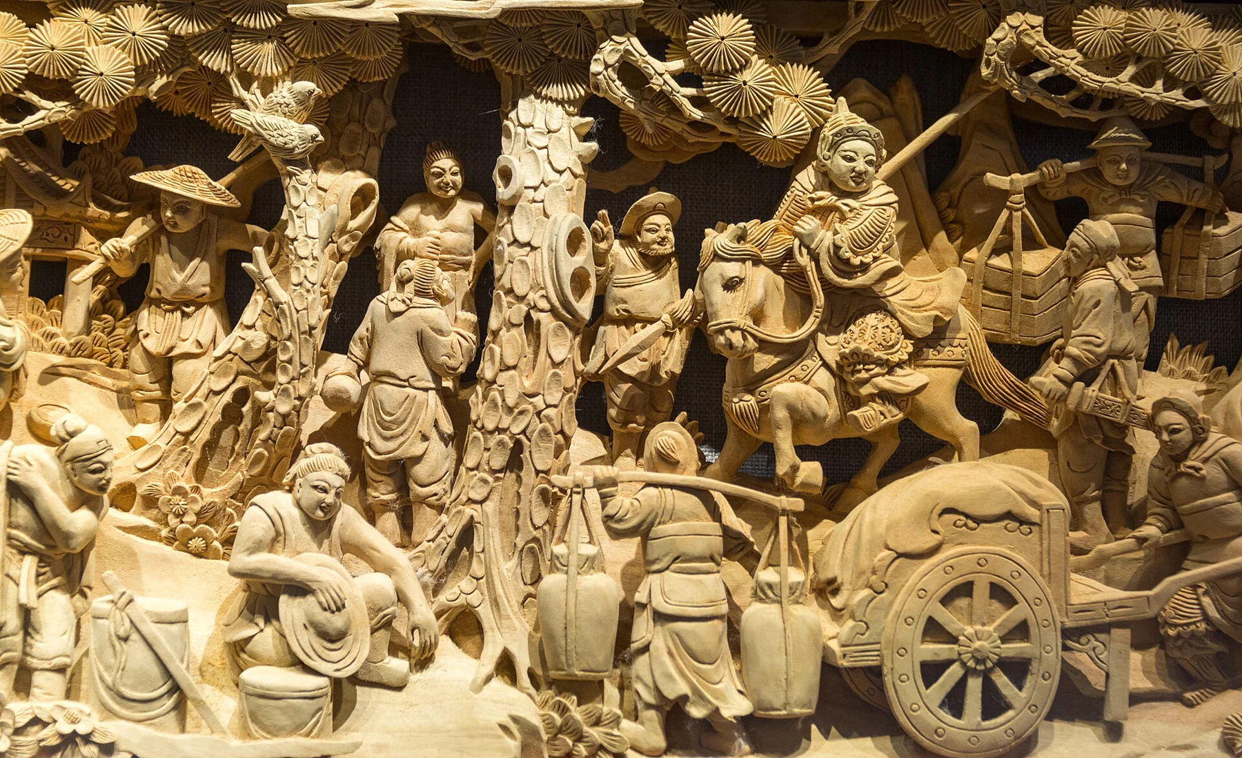 Carved narrative scene with figures, trees and a wheeled vehicle.