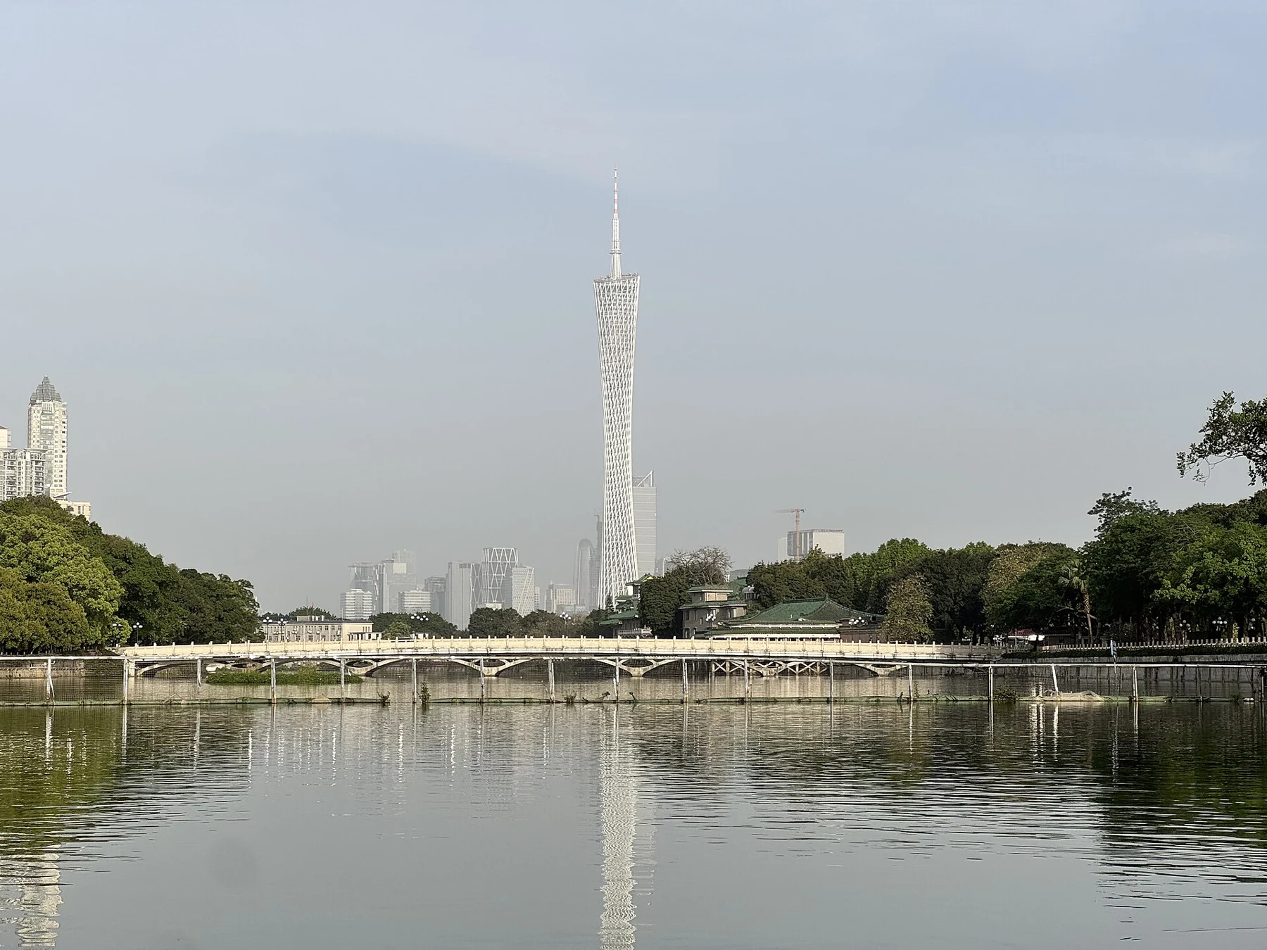 Distant tower seen beyond a lake, trees and a hazy city sky.