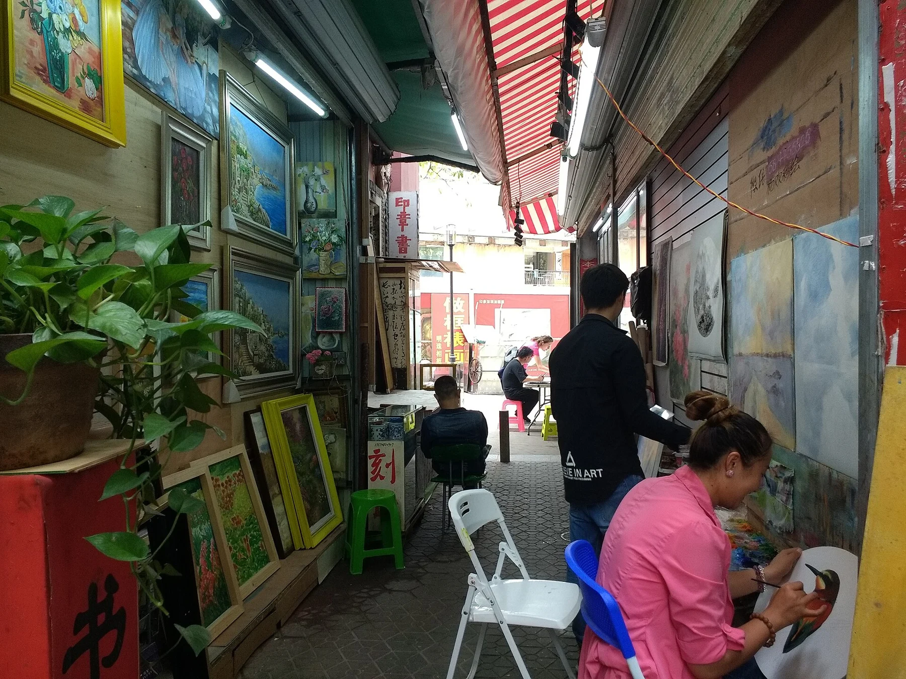 People painting beside displayed pictures and stools under striped awnings in a narrow lane