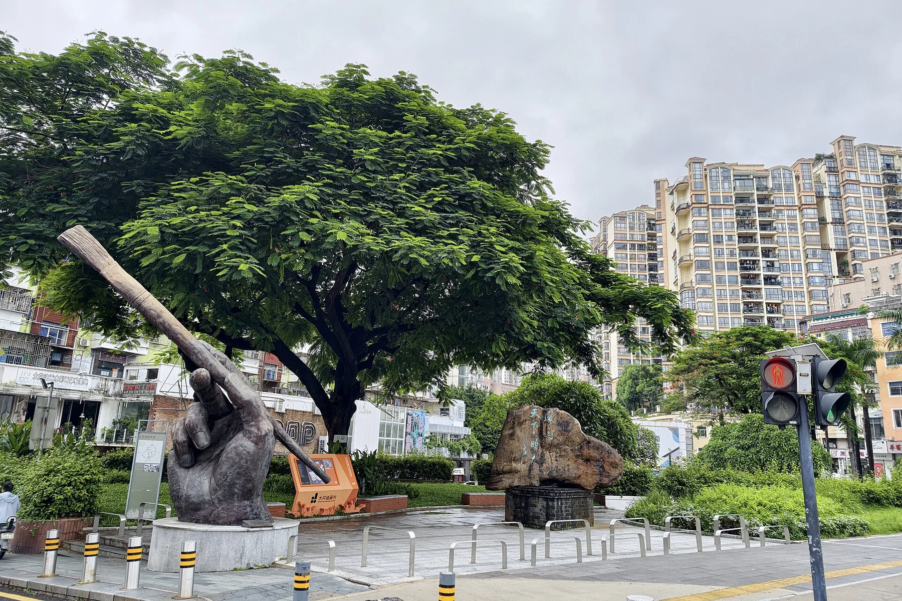 Large hand holding a brush beside a stone sign, trees, street signals and surrounding buildings