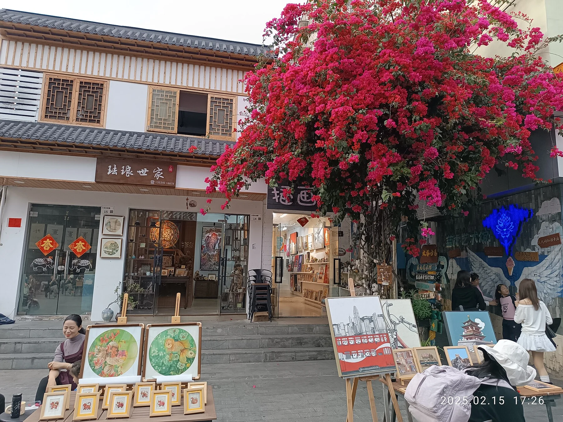 Pink-red bougainvillea, tiled eaves, shop doors, displayed pictures and people on a Dafen lane
