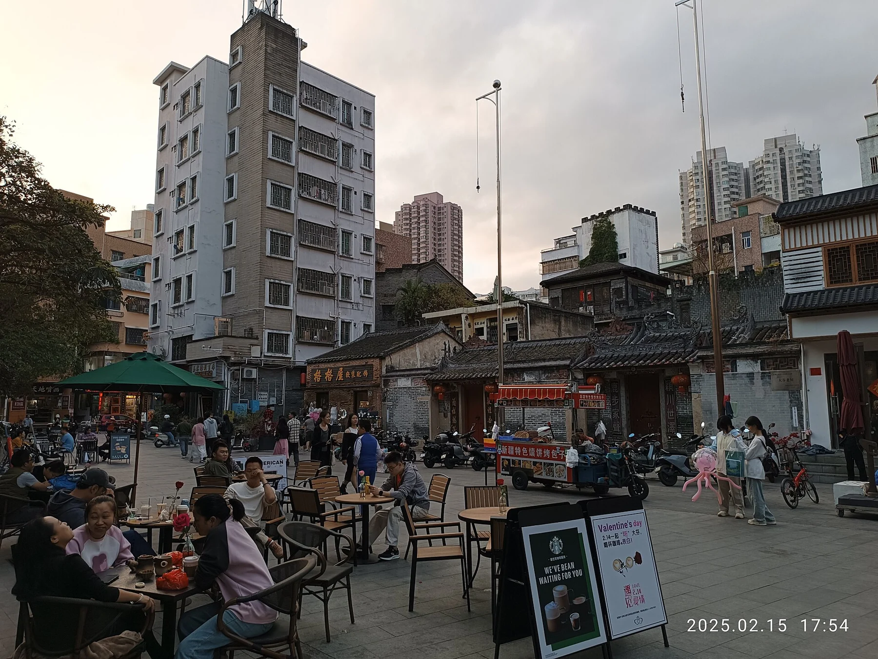 People sitting at outdoor tables before low tiled-roof buildings and taller housing