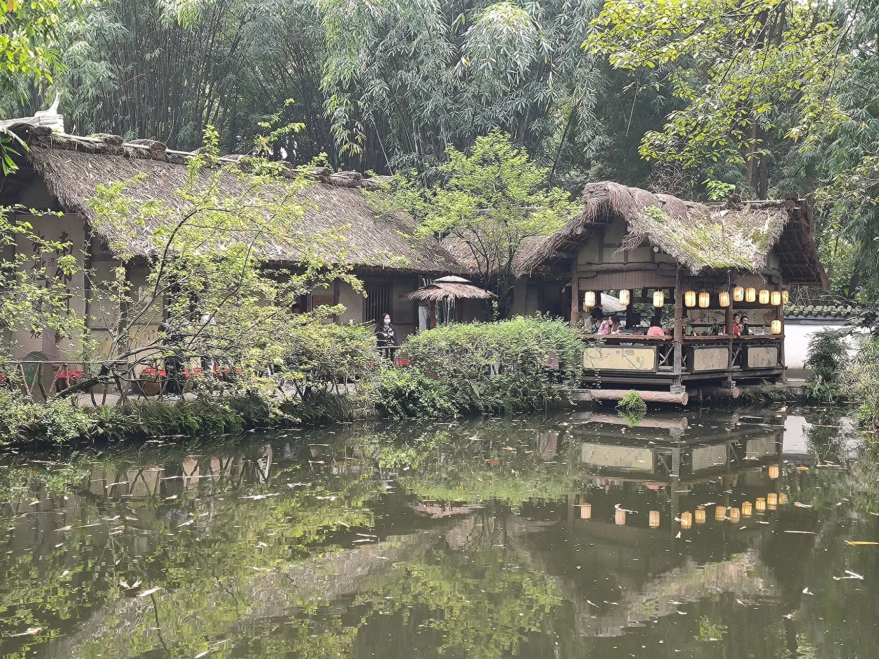 People, lanterns and a counter in thatched waterside buildings; April 2023. Not identified as the residential reconstruction.