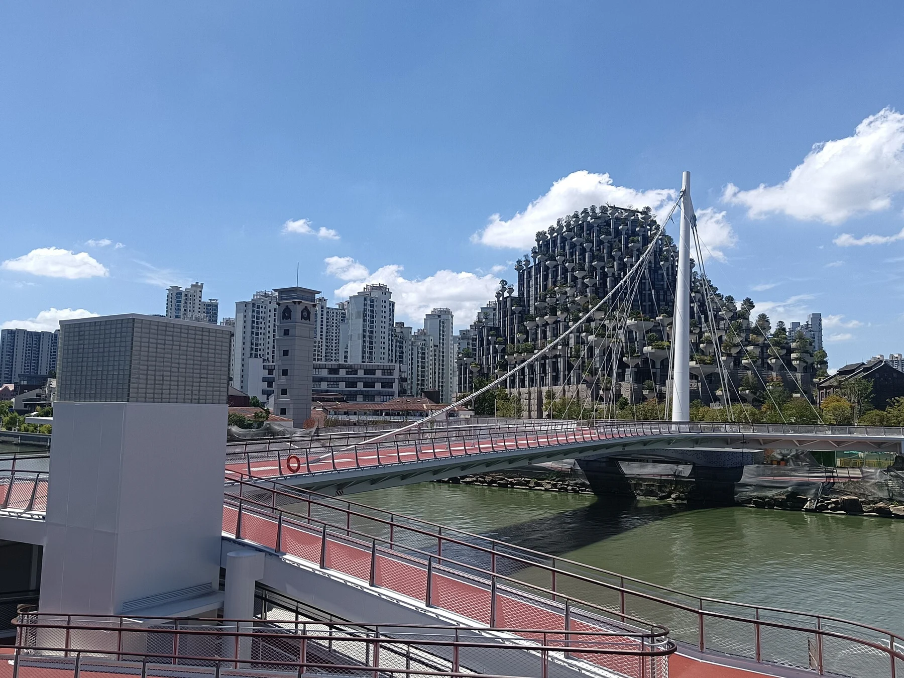 A red-deck pedestrian suspension bridge crossing Suzhou Creek in front of a planted building complex.