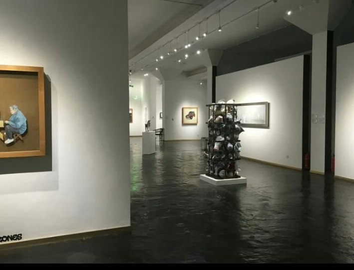 A gallery room with white walls, a dark floor, framed works and a sculpture in the centre.
