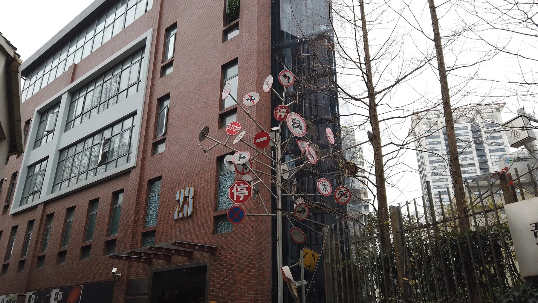 A brick-and-glass factory building marked 23 beside a pole carrying many round signs.