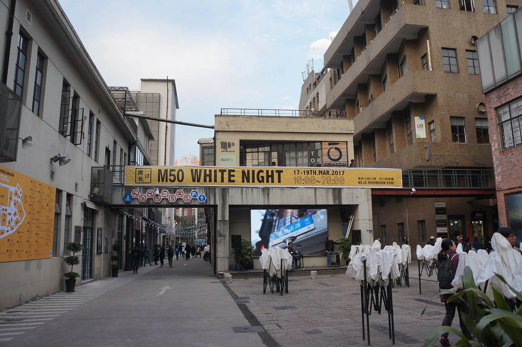 An industrial courtyard with an event banner, plants and white shirt-like forms on stands.
