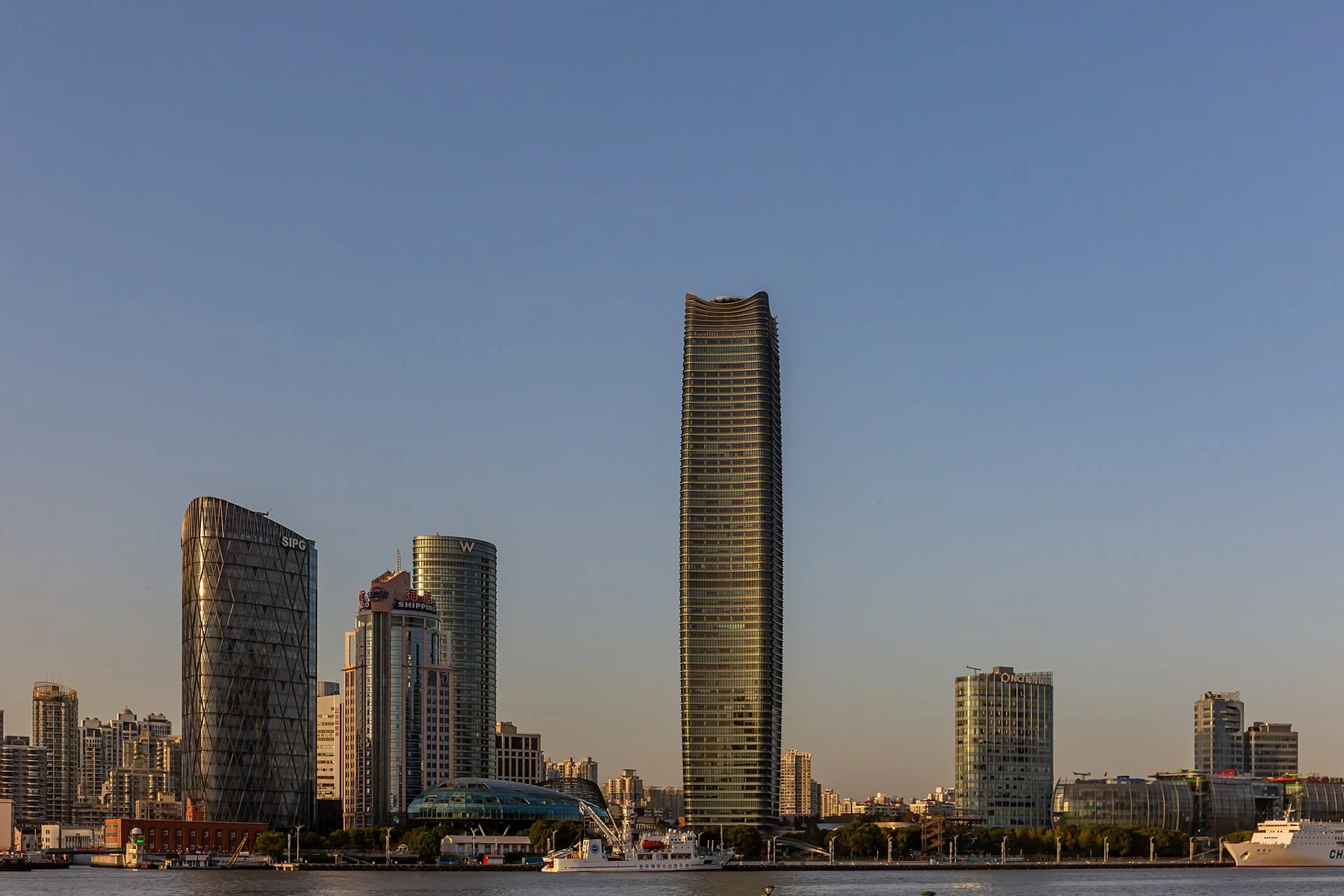 A group of riverfront towers with the tallest building catching warm light above a low green embankment.
