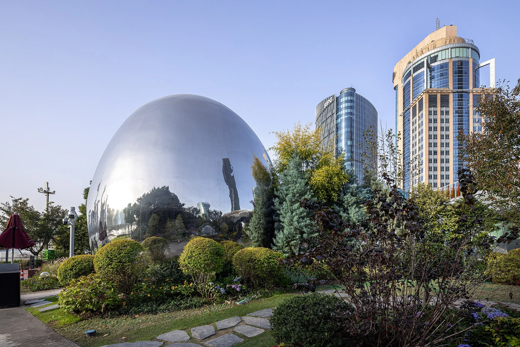 A large mirrored oval form among trees and shrubs with towers behind.