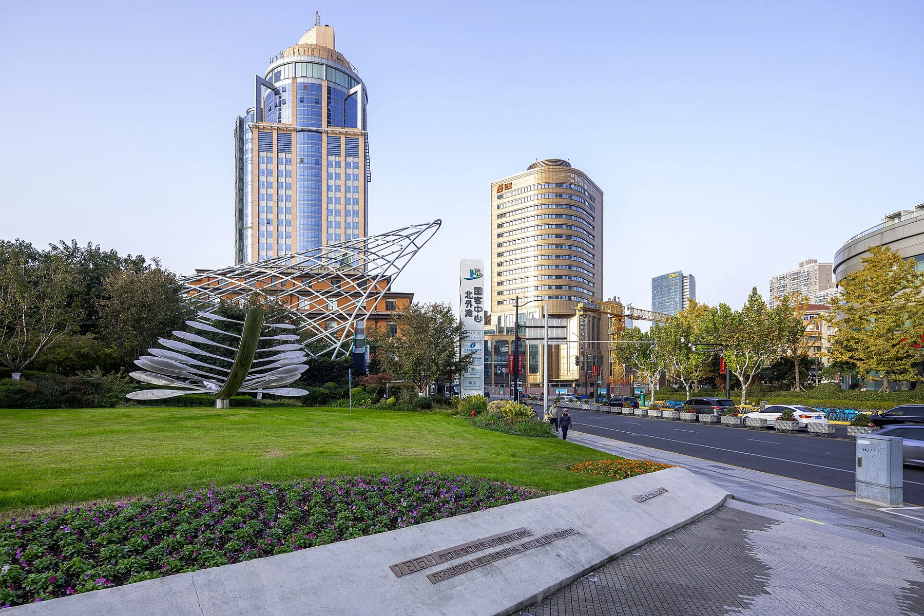 A broad paved approach, green planting and a curved metal sculpture beside city buildings.