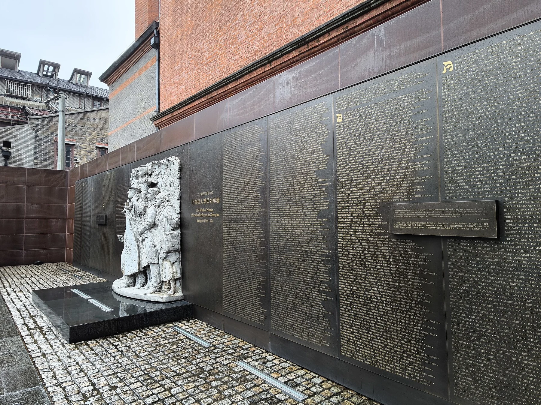 Dense engraved name panels beside a pale group sculpture in a narrow brick-lined courtyard.
