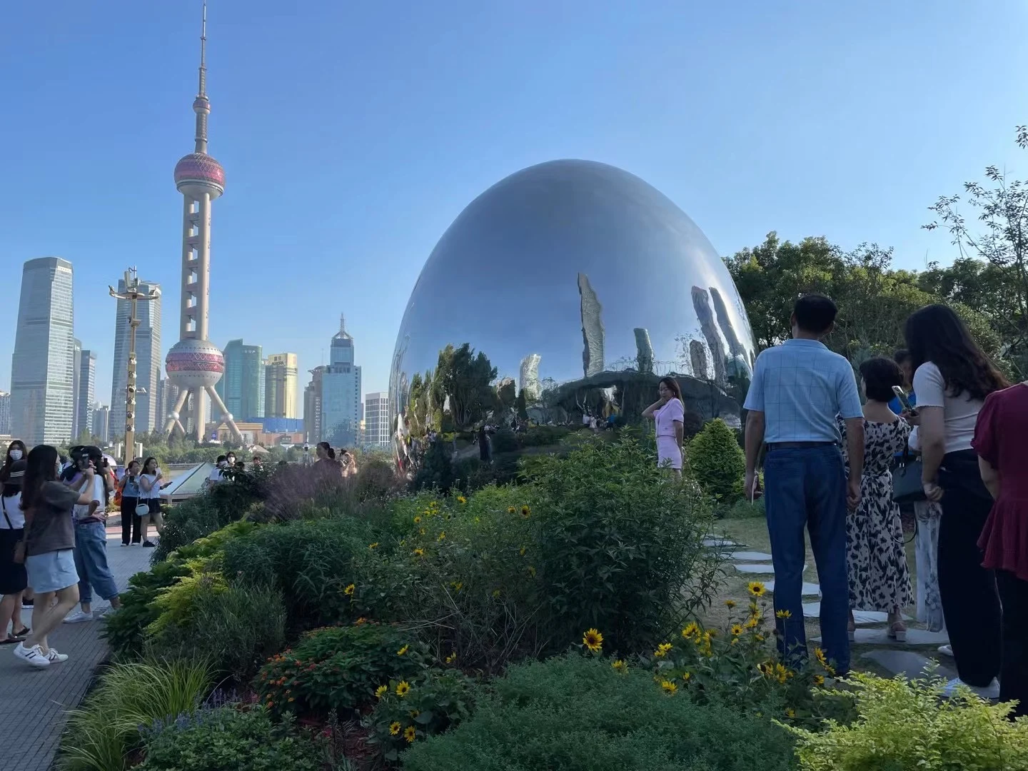 People on a narrow planted path near a large mirrored oval structure with the Oriental Pearl beyond.