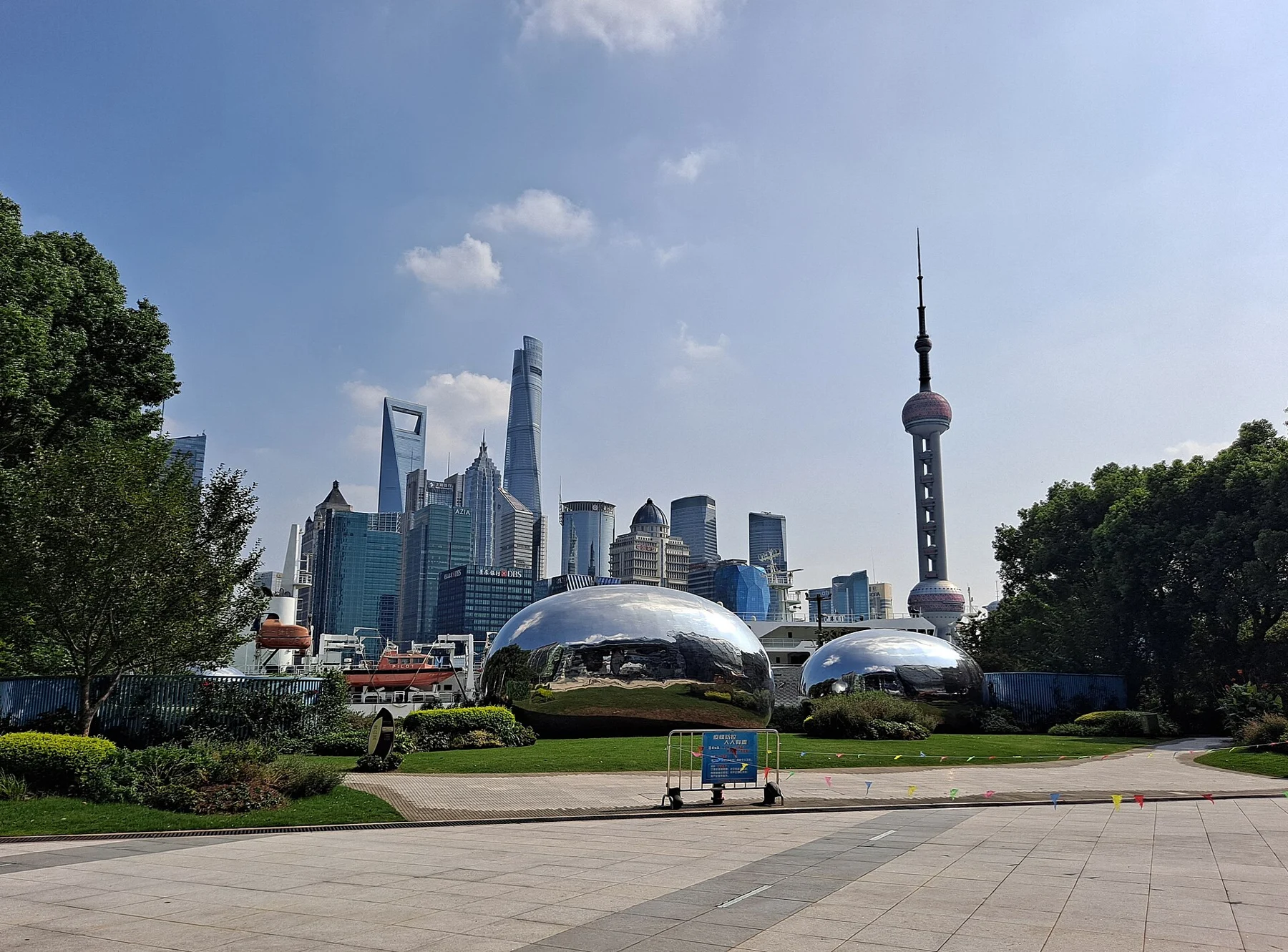 Broad paving and grass beside reflective rounded forms, with the financial-district skyline across the river.