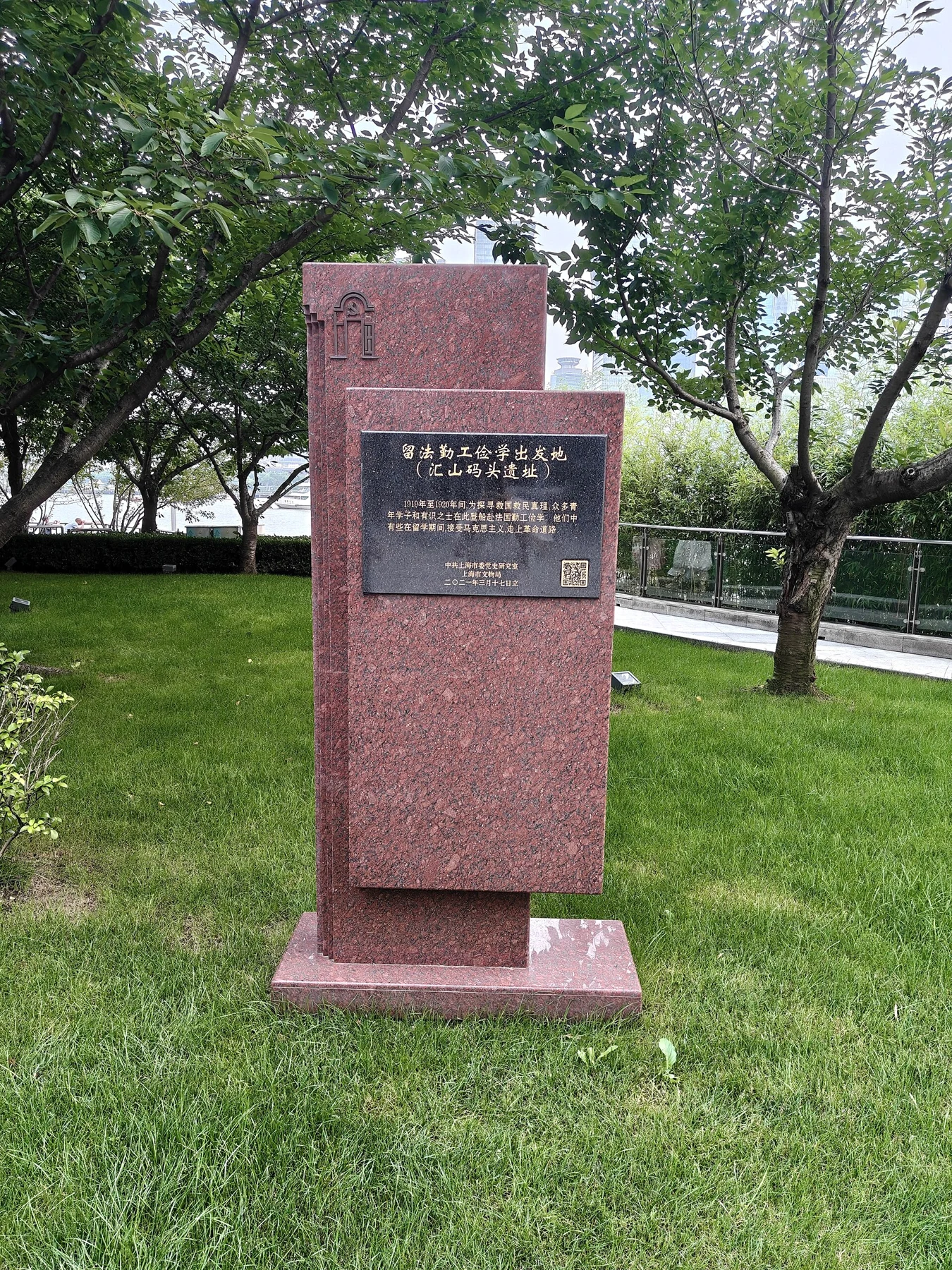 A tall red stone marker with an inscription panel beside trees and a path.