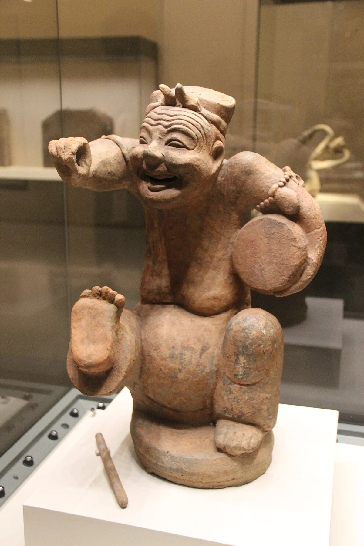A lively seated pottery performer holding a small drum and raising one arm.