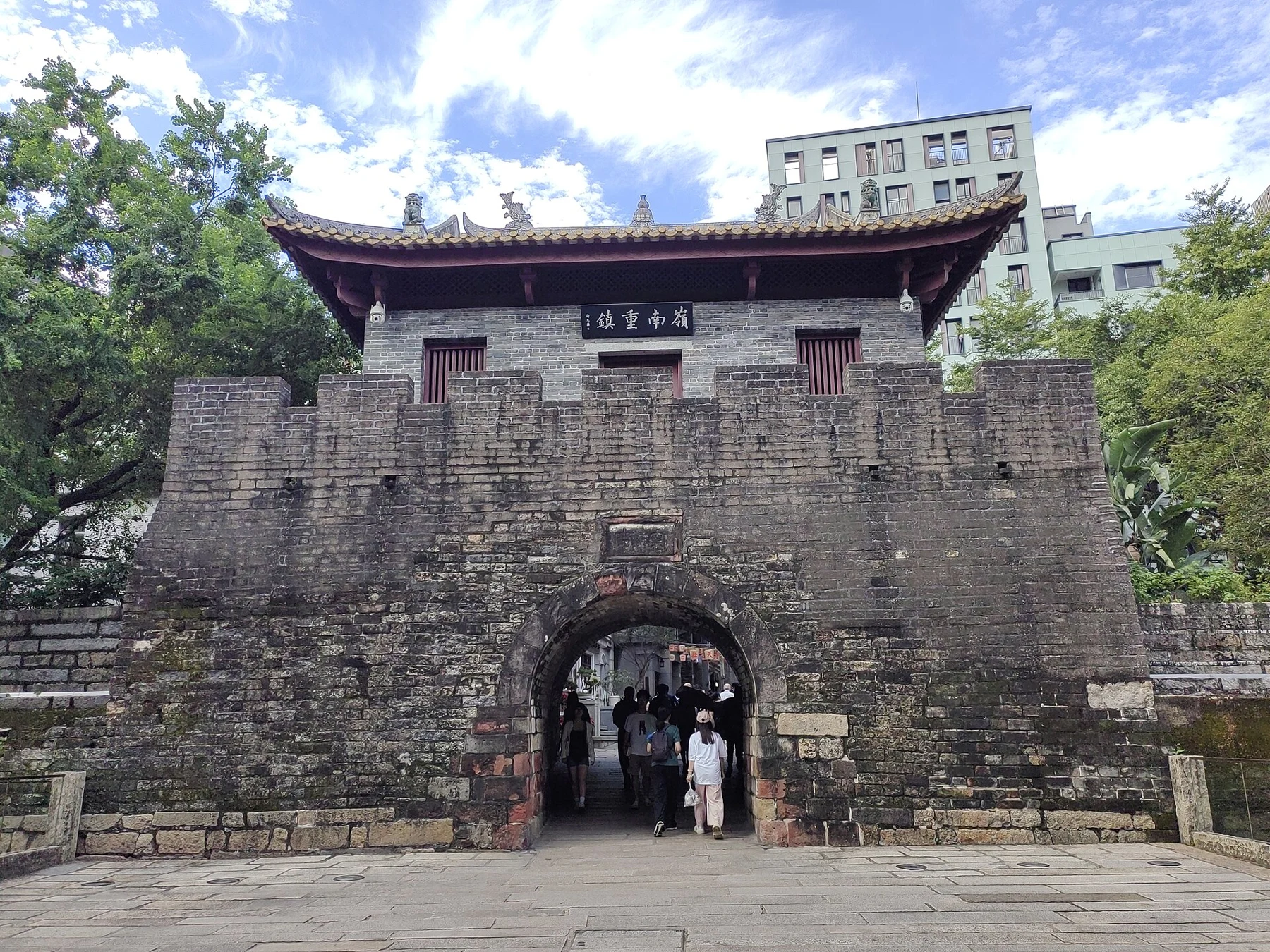 Brick south gate with a stone arch, red-windowed upper building and surrounding modern buildings
