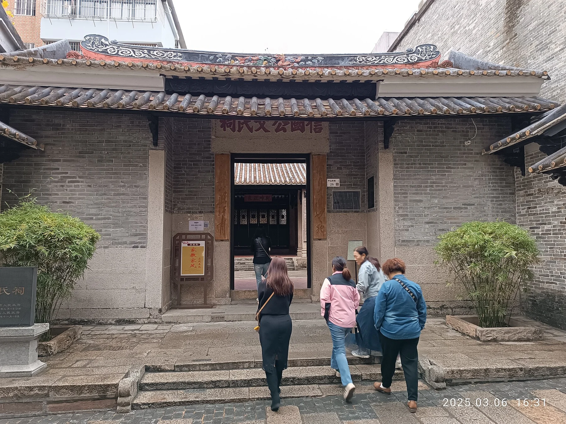 Grey-brick ancestral hall entrance with carved roof decoration and visitors on the steps
