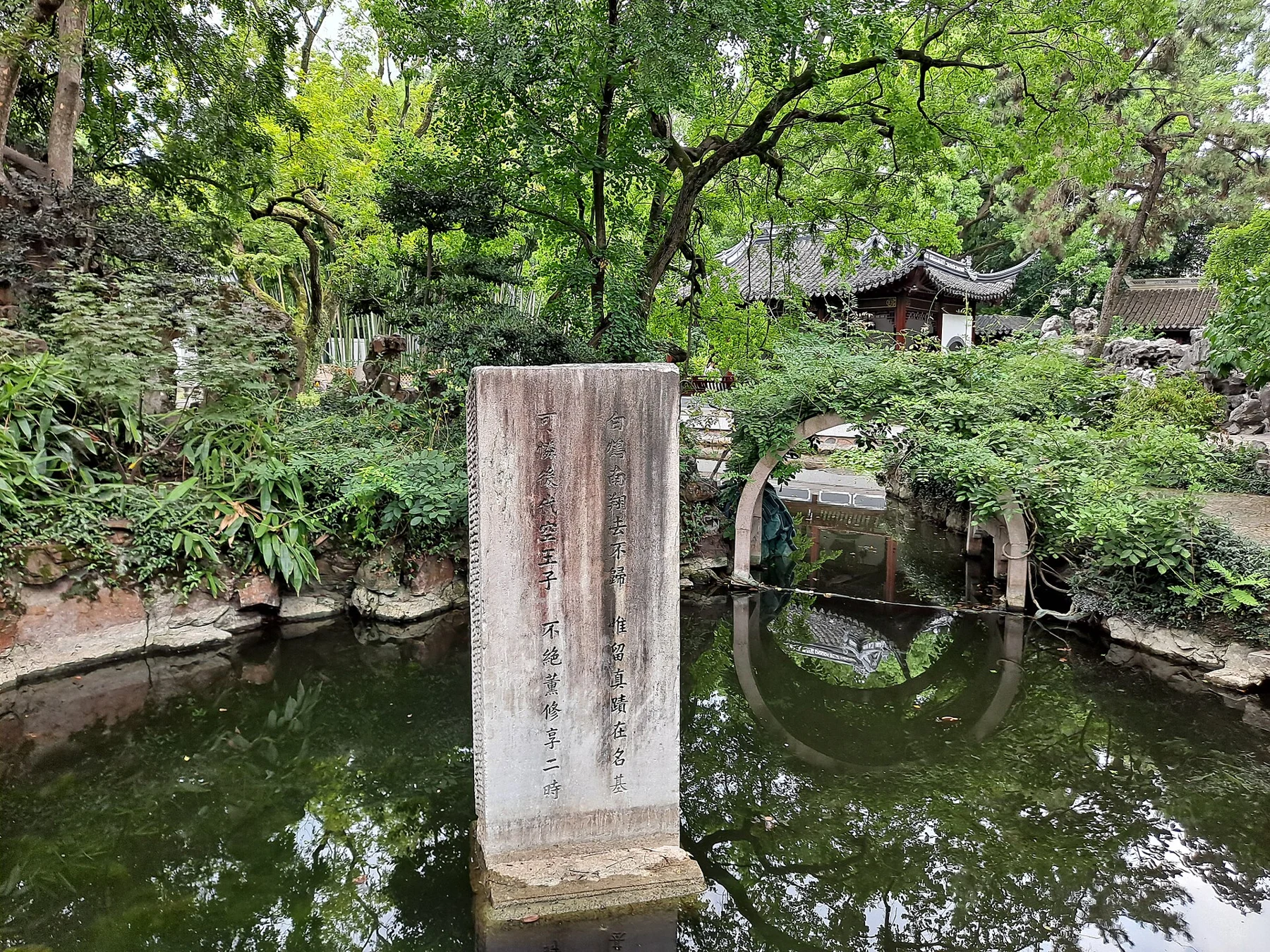An inscribed upright stone in water, with an arched bridge and planting behind.