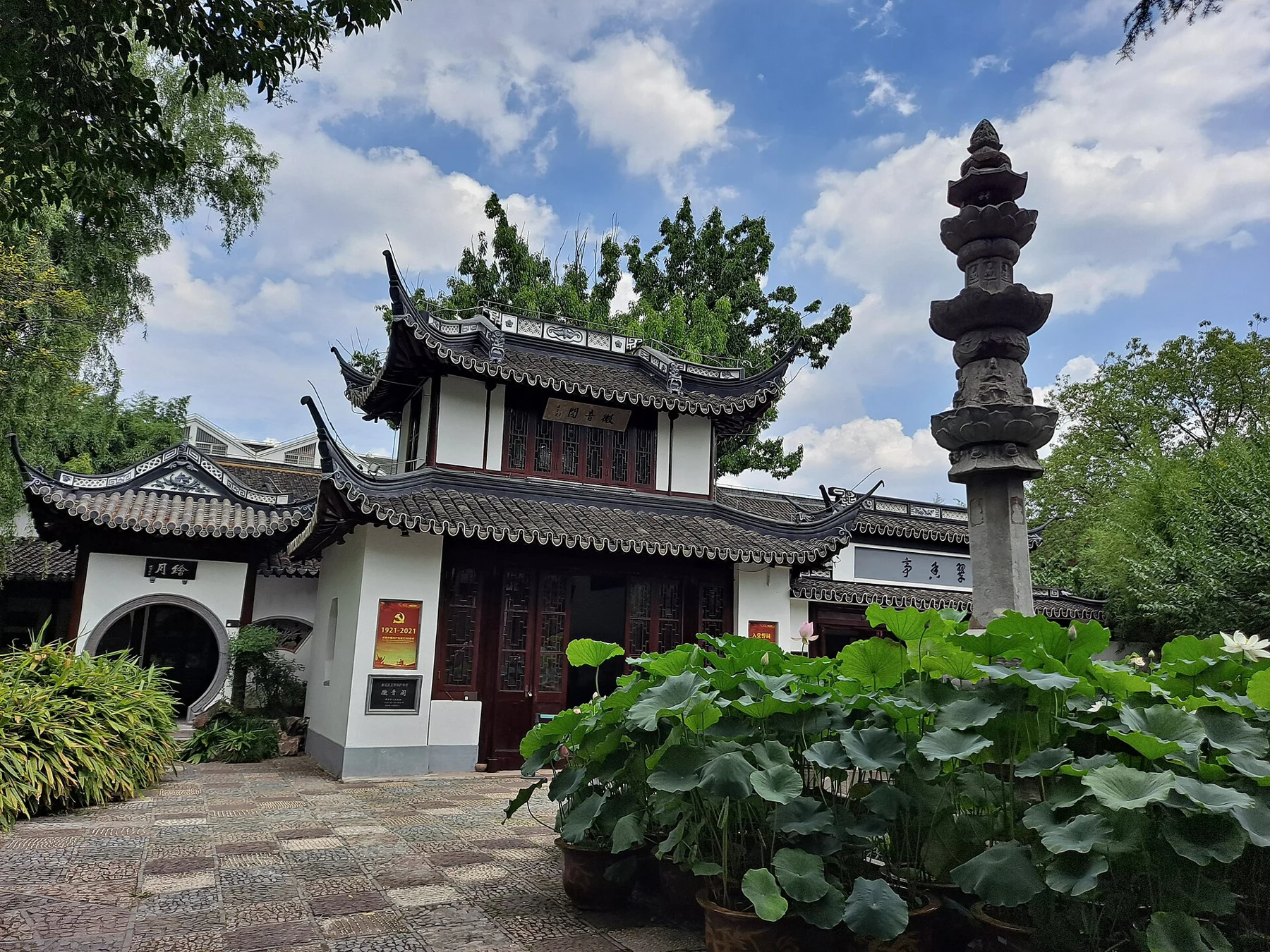 A tiered stone pillar beside a small white-walled building and lotus leaves.