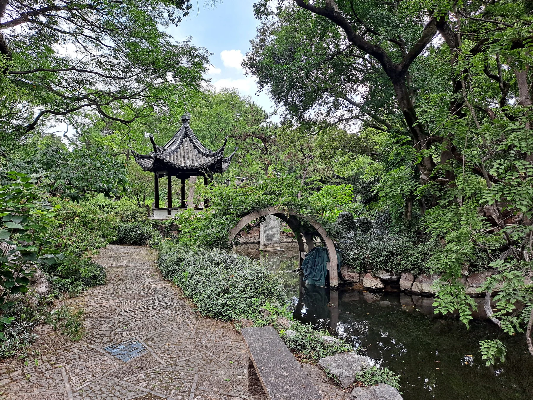 A curved path beside an arched stone bridge and a small open pavilion among trees.