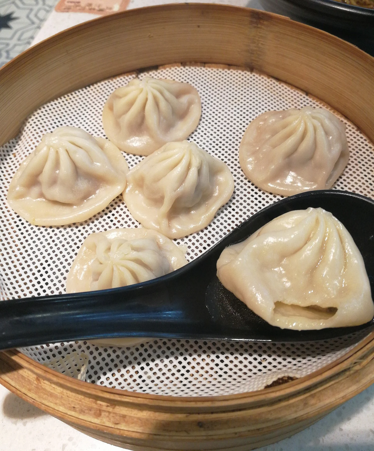 A bamboo basket of pleated soup dumplings, with one resting on a black spoon.