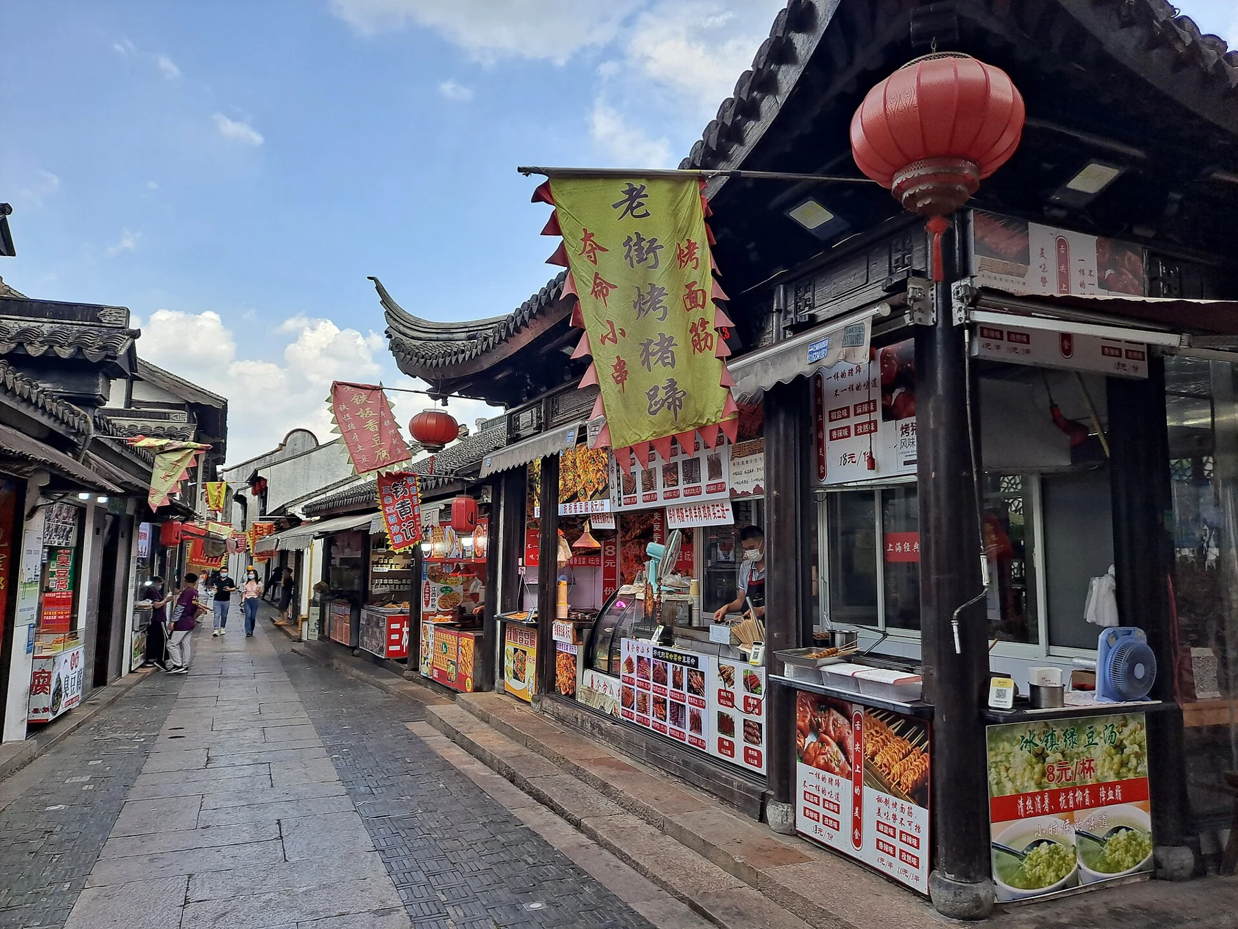 A paved lane lined by low shopfronts, hanging lanterns and traditional tiled roofs.