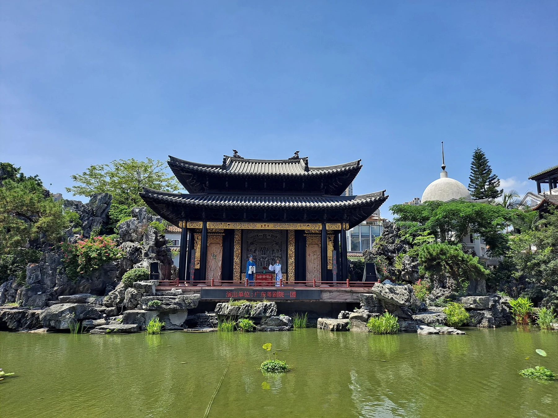 Frontal view of the black-and-gold stage reflected in a green garden pond