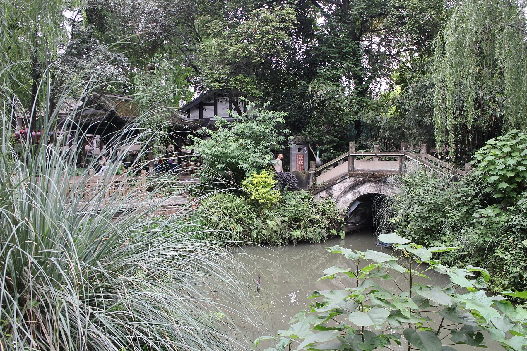 A stone arch bridge and timber pavilion reflected in a narrow planted watercourse.