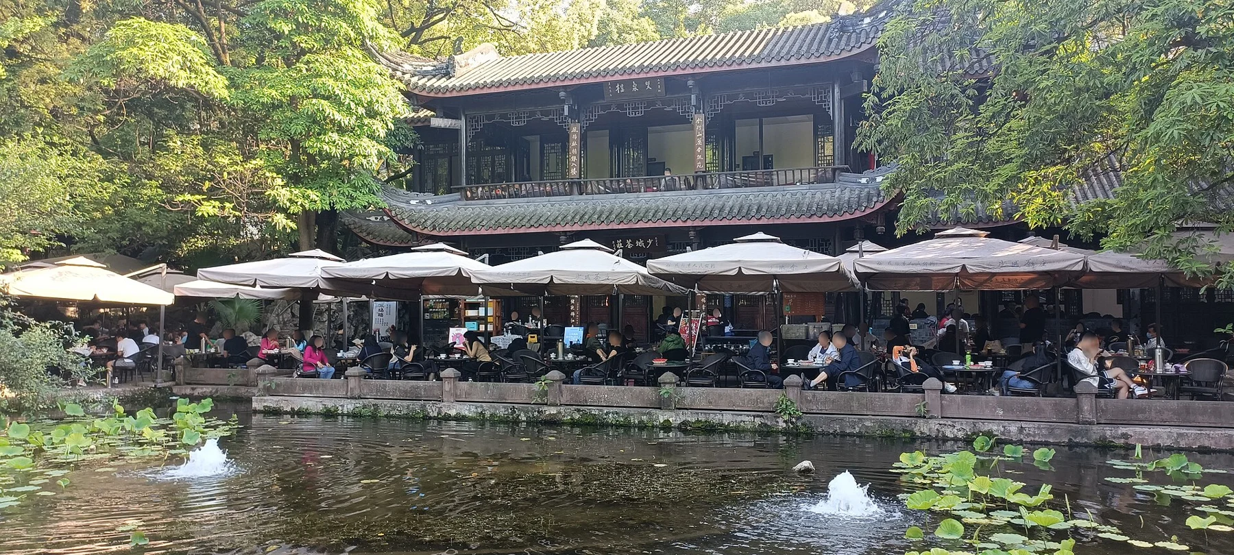 A two-storey tiled-roof teahouse behind parasols and tables beside a pond.