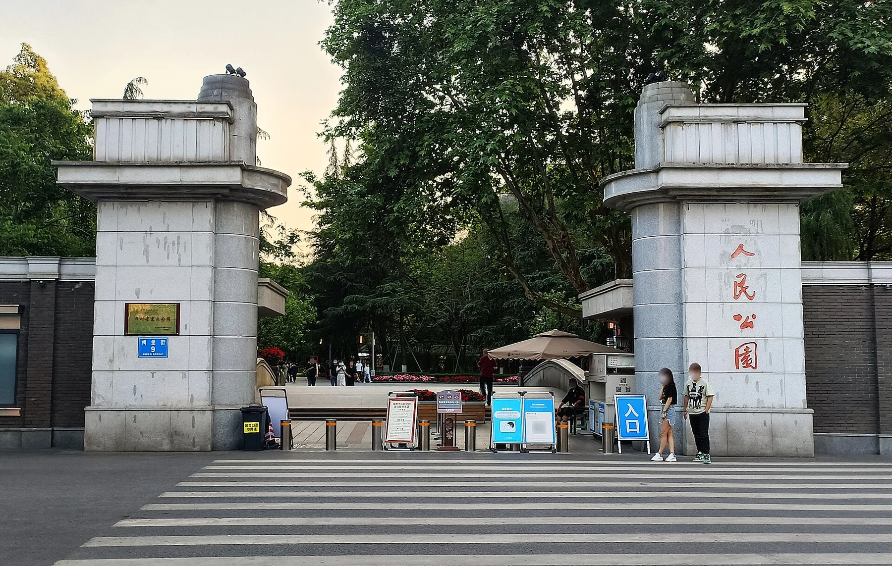 Two pale gate posts with People’s Park characters, trees beyond and entry barriers in a 2022 photograph.