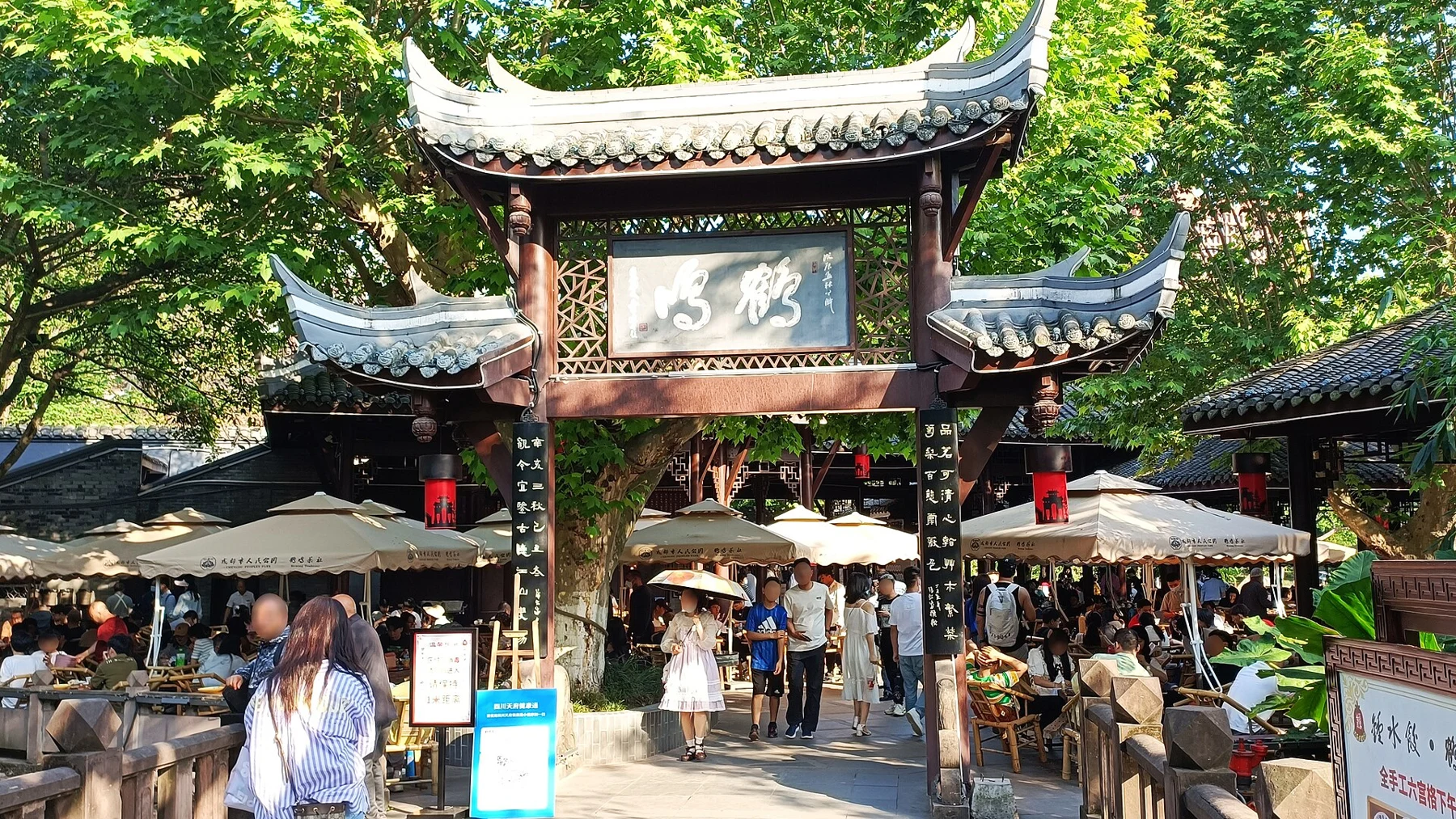 A timber gateway bearing Heming characters, with parasols, trees and seated visitors beyond.
