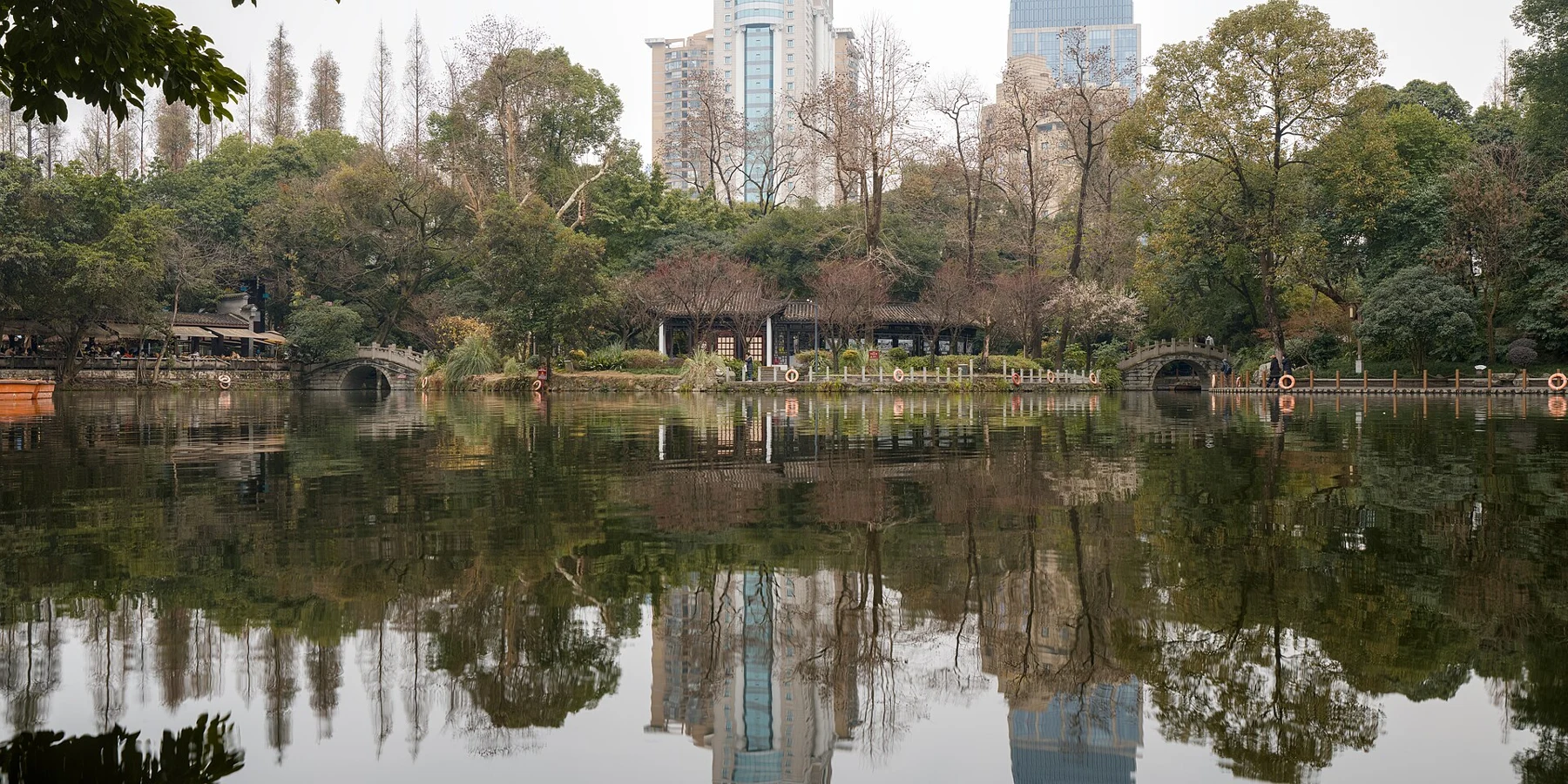 A broad park lake reflecting trees and city buildings, with stone bridges on the far side.
