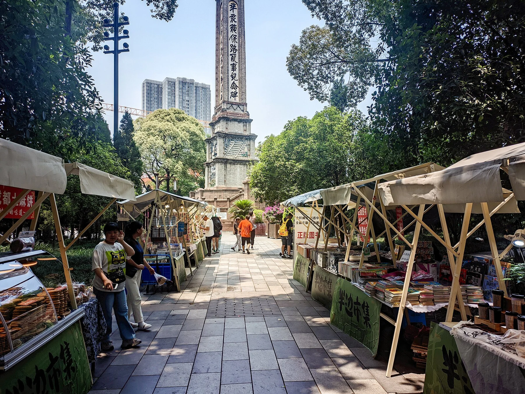 A pedestrian lane between stalls beneath canopies, leading toward a tall memorial tower.