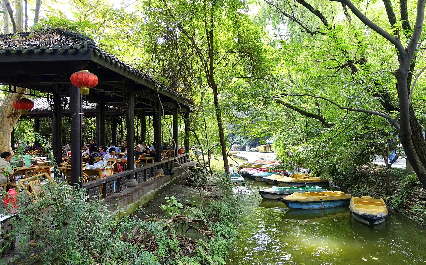 Seated tea drinkers beneath a roof beside water and several small moored boats.