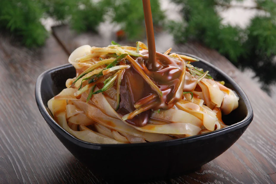 Broad cold noodles with cucumber and a brown sesame-paste dressing being poured over them.