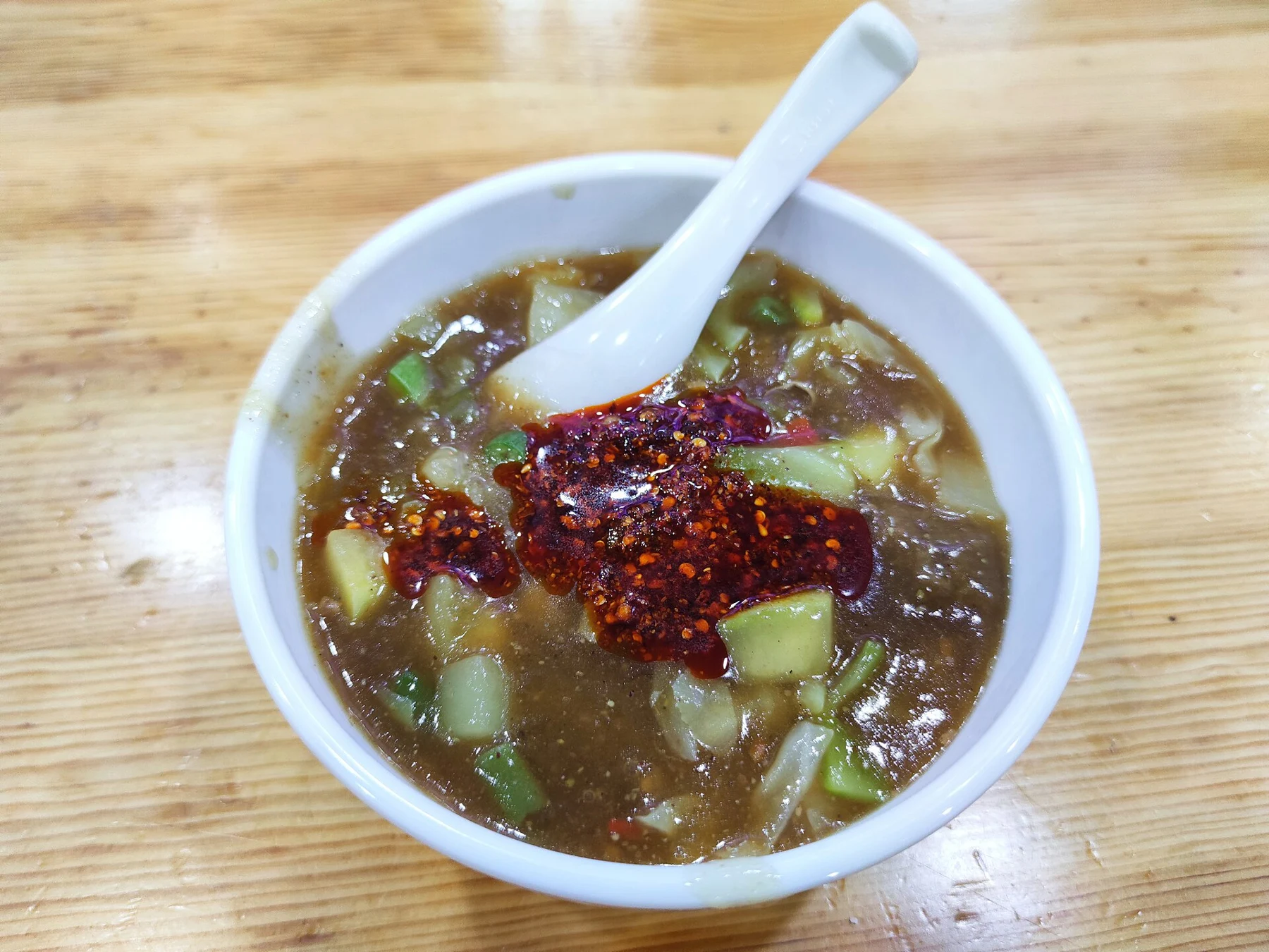 A spoon in a thick meatball-and-vegetable soup with red chilli oil on top.