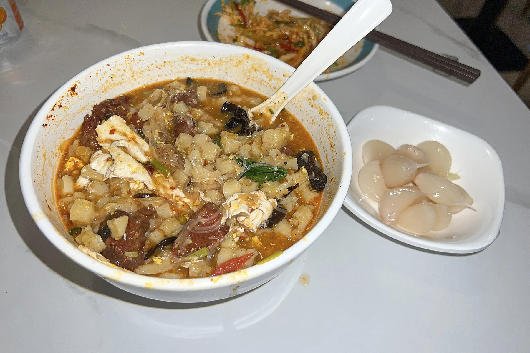Bread pieces, meat and vegetables in a sauced bowl, beside a plate of garlic.