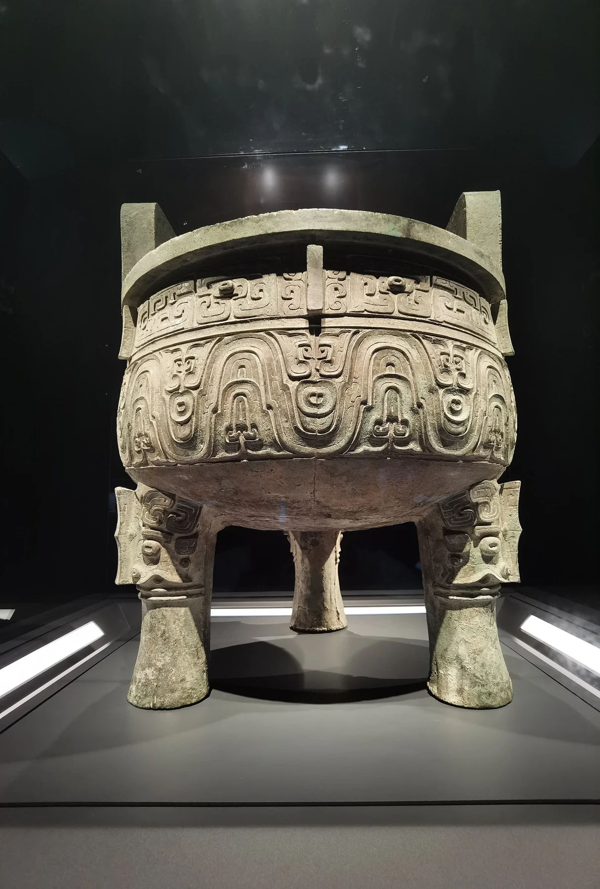 A large bronze tripod vessel with two upright handles, broad decorative bands and three heavy legs in a lit case.