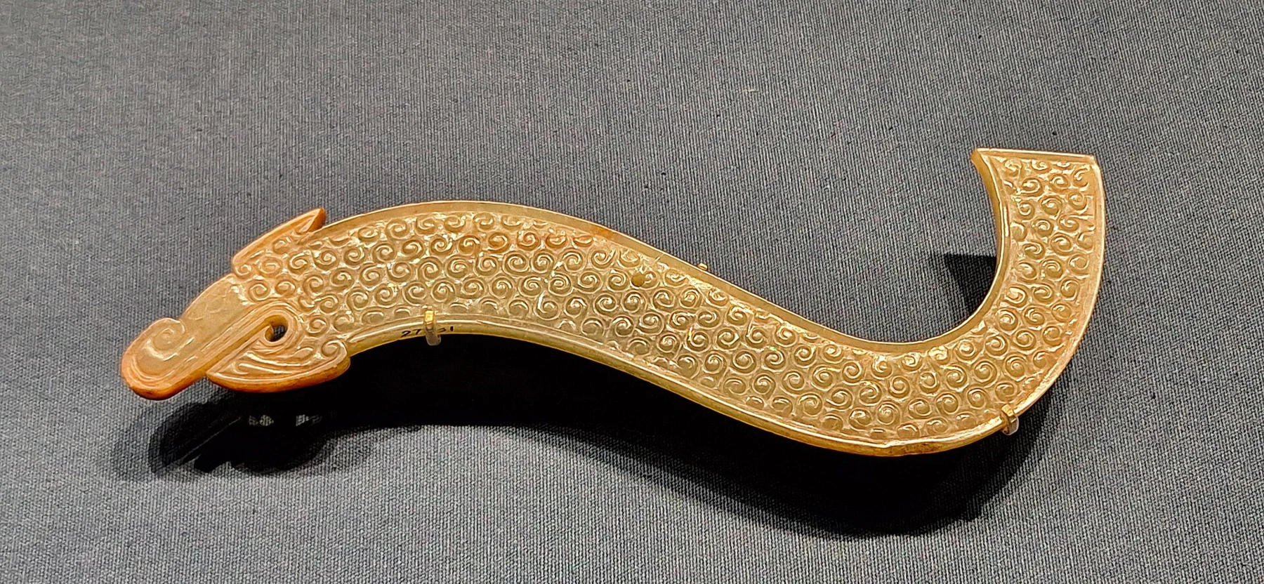 A curved yellow-brown carved ornament with an animal head at one end and dense small patterns across its surface.