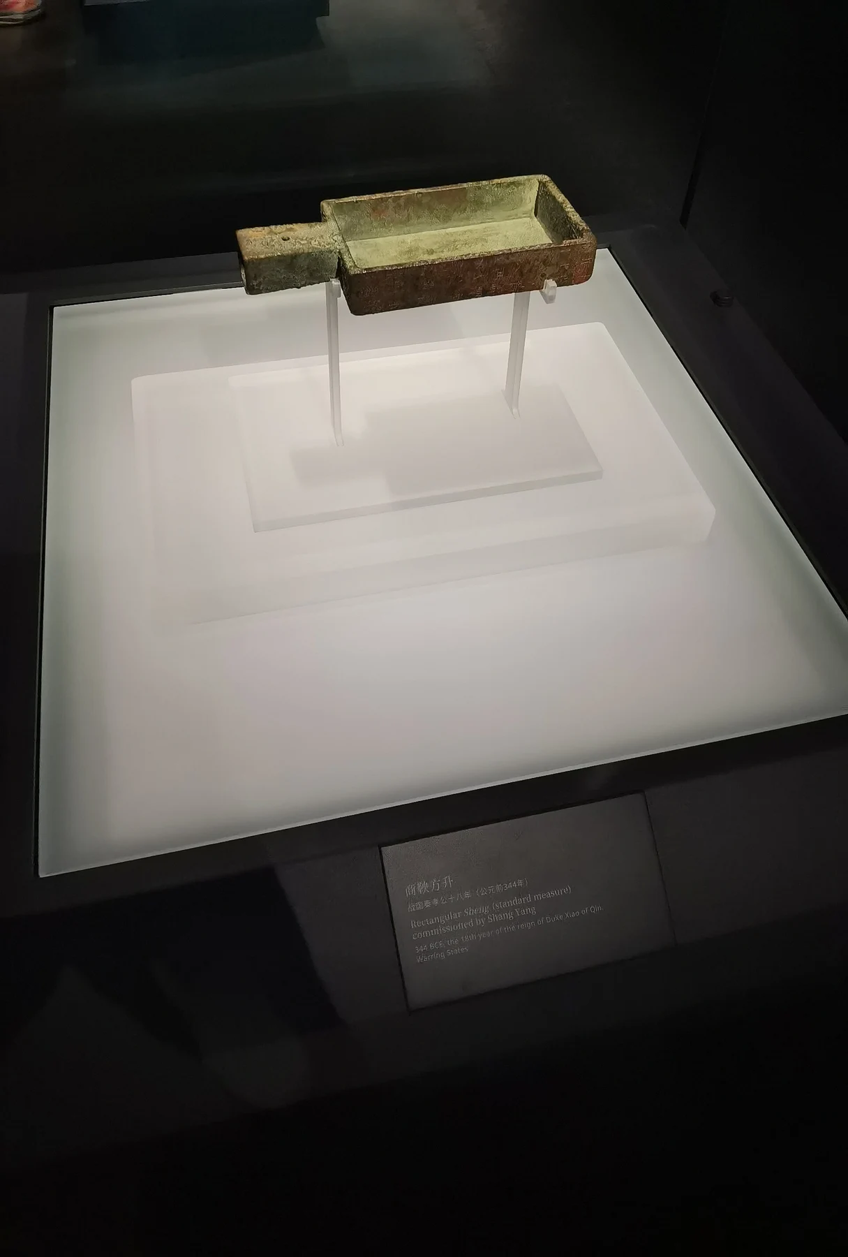 A small rectangular bronze measuring vessel and handle displayed on a pale support.