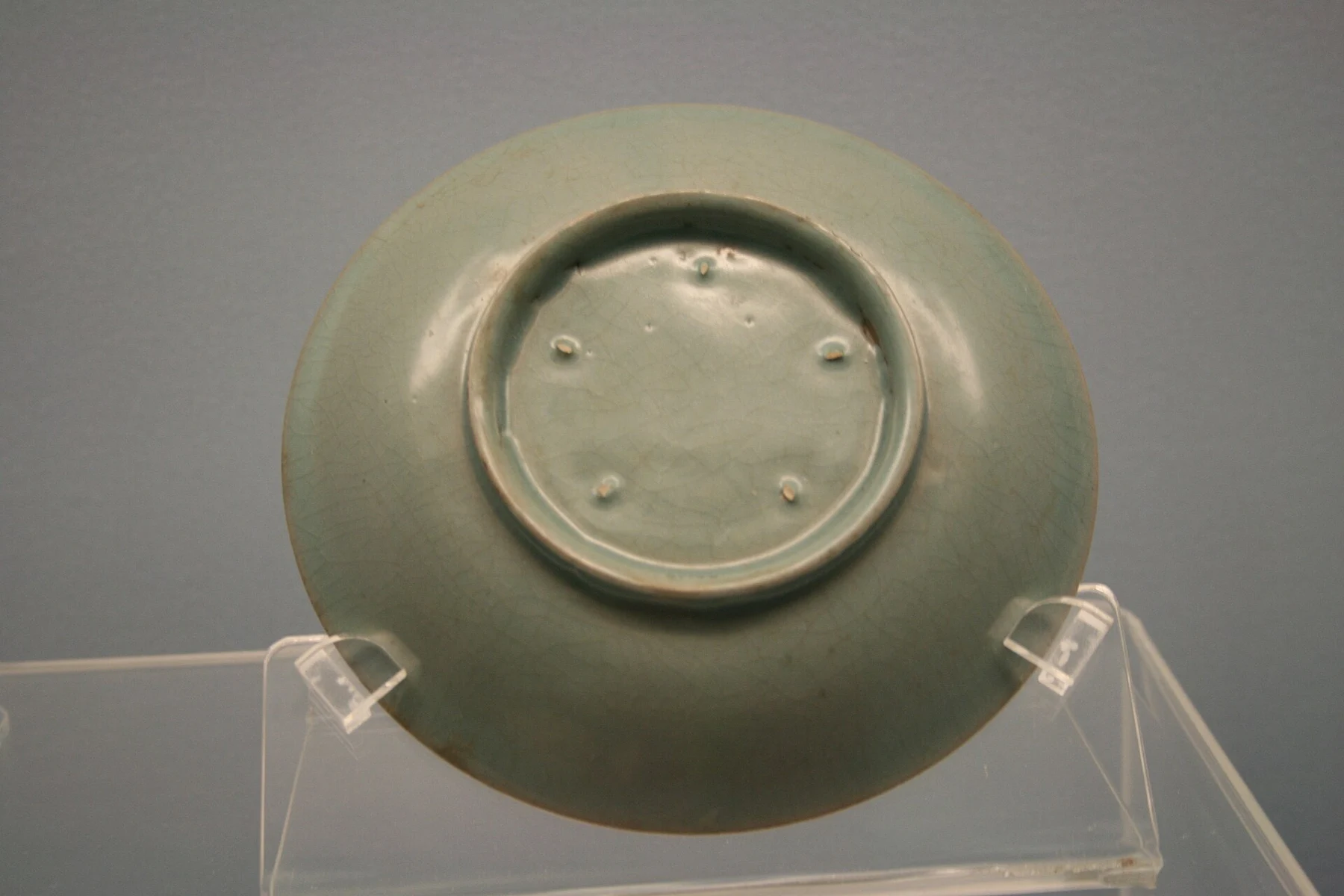 The underside of a glazed dish on a clear stand, showing five small firing-support marks.
