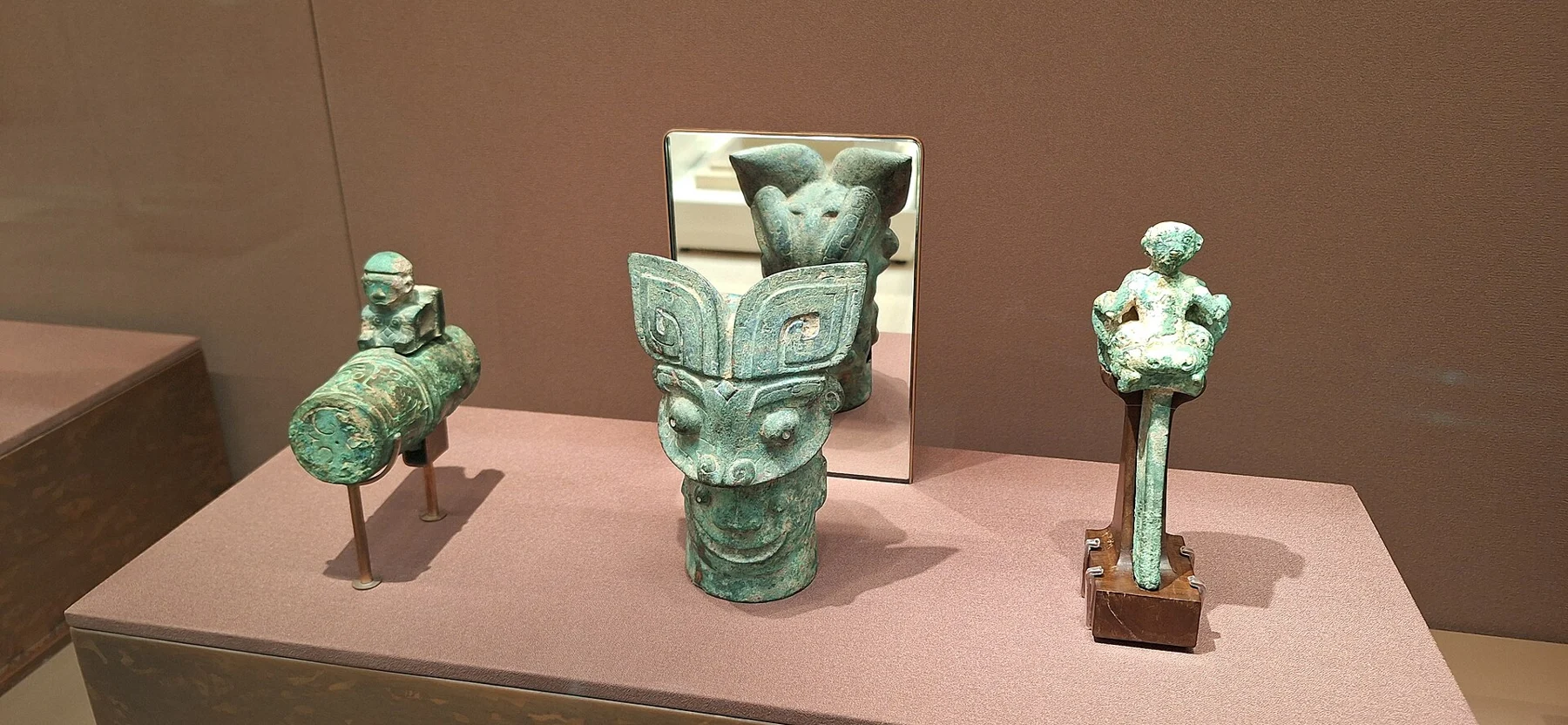 Three small green-patinated bronze objects, including a tall mask-like central form, displayed on a plinth.