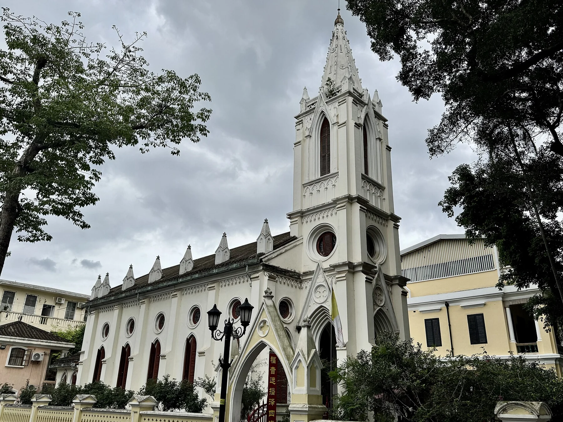 Our Lady of Lourdes church with a pale tower and pointed doorway beneath trees.