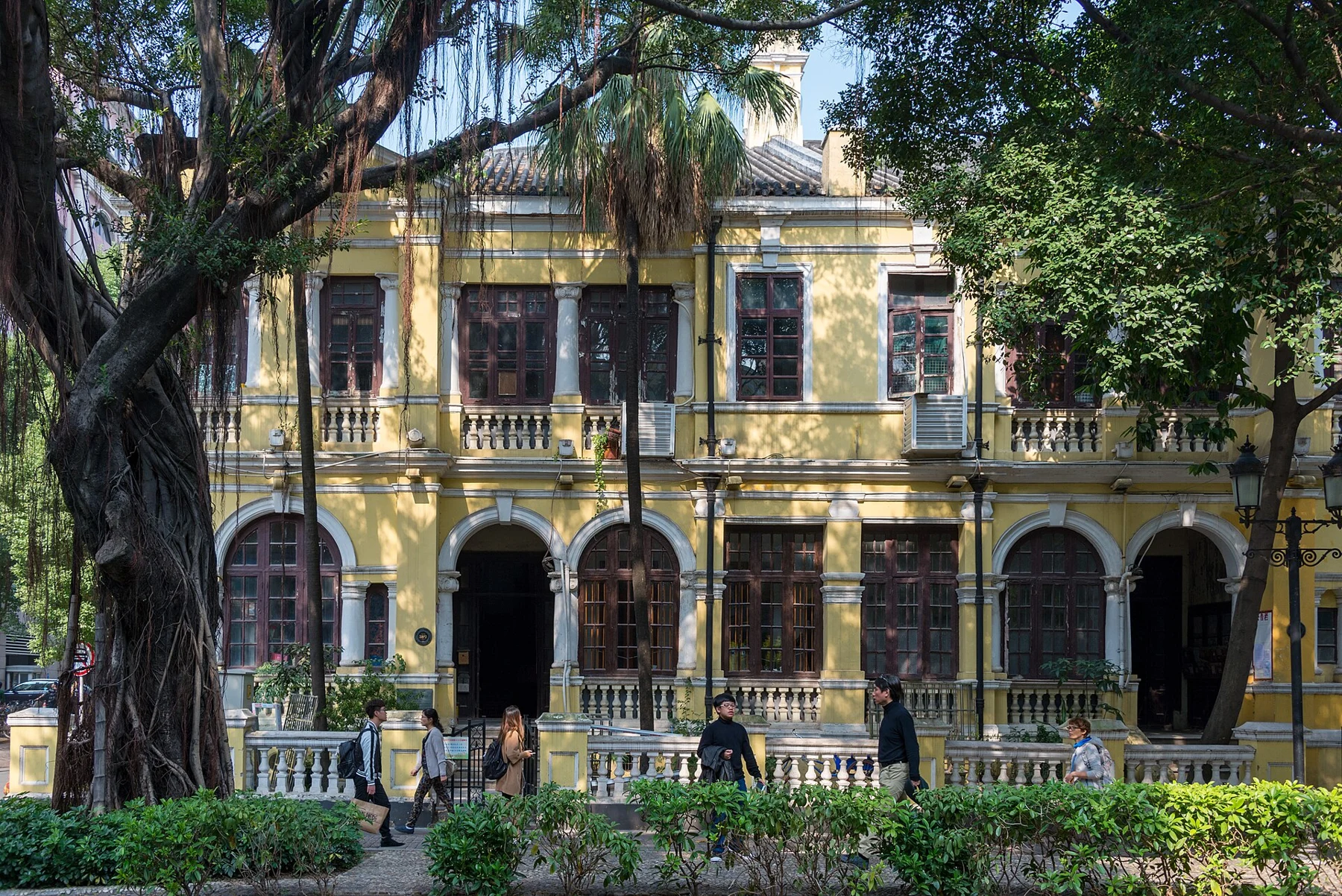 Yellow building with arched lower openings, railings and trees in front.