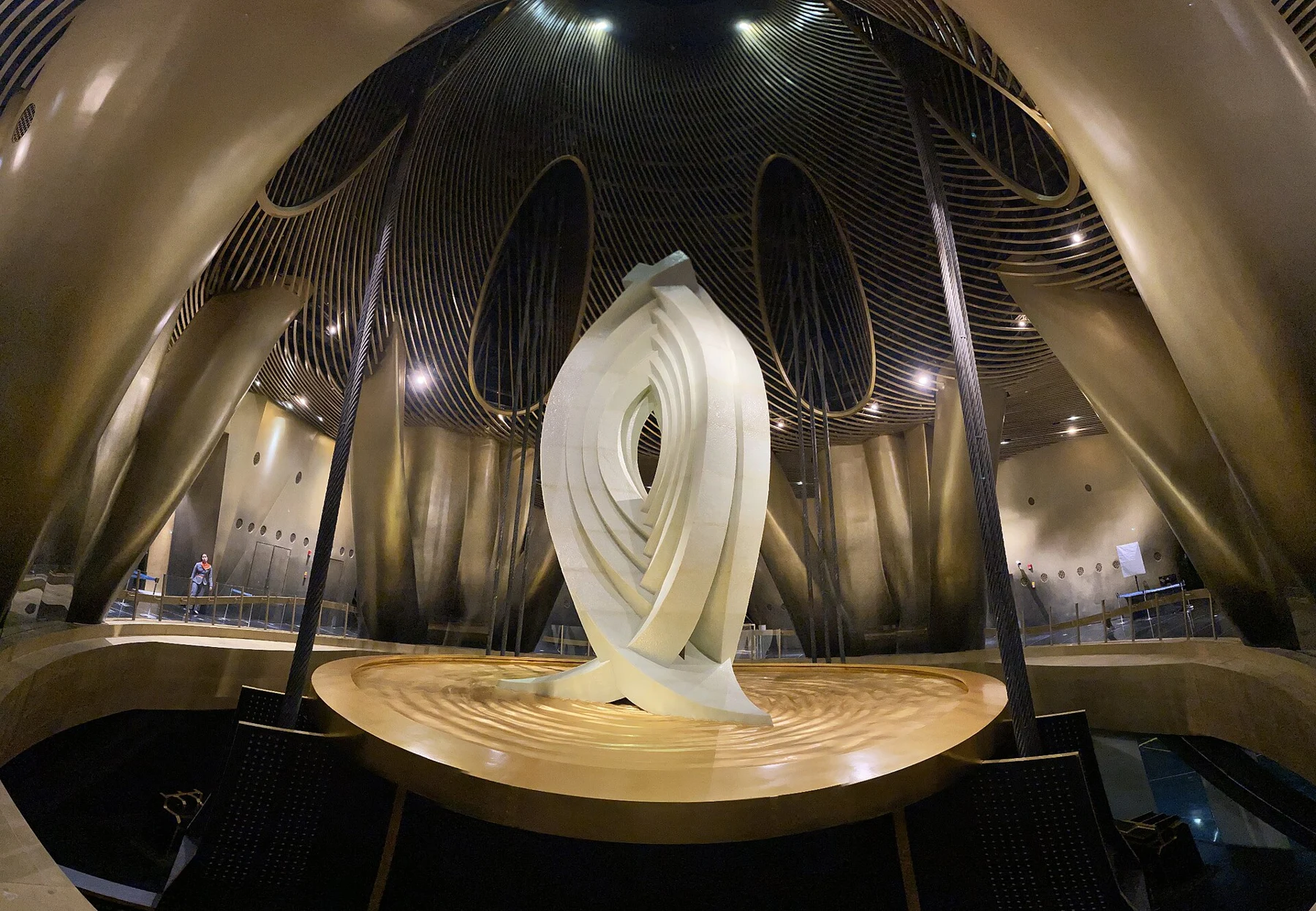 White eye-like sculptural installation above a round platform in a dark interior.