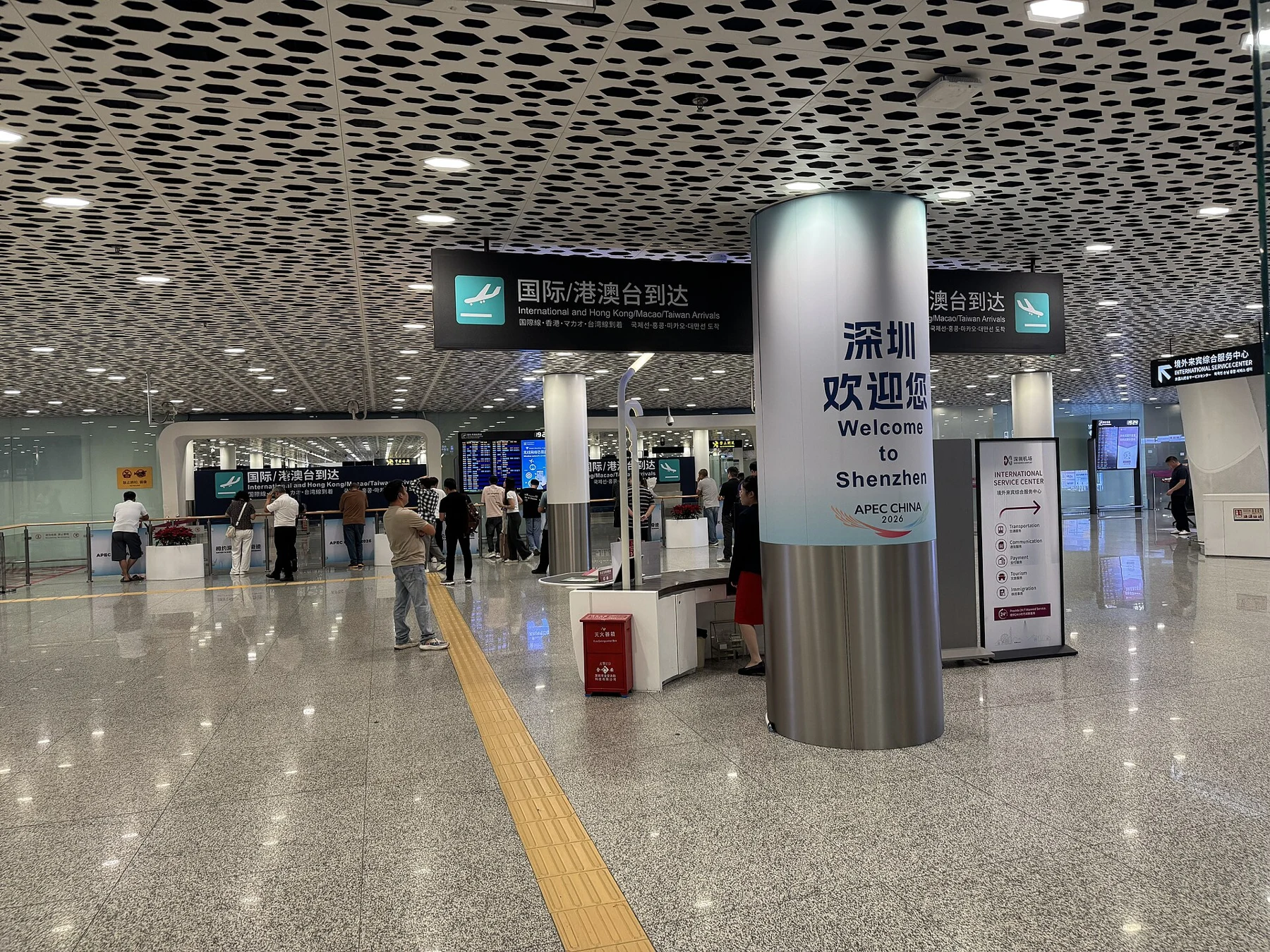 Airport arrival hall with a tactile floor strip, overhead signs and a welcome pillar.