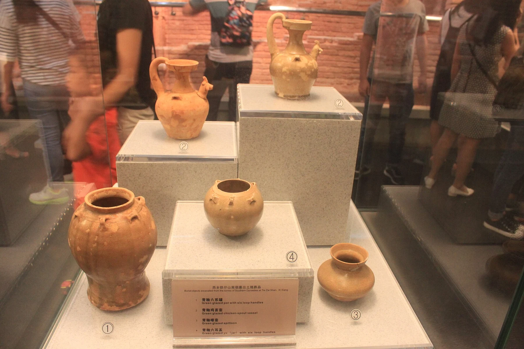 Two vessels with chicken-shaped spouts, jars with loop handles and a wide-mouthed spittoon behind glass