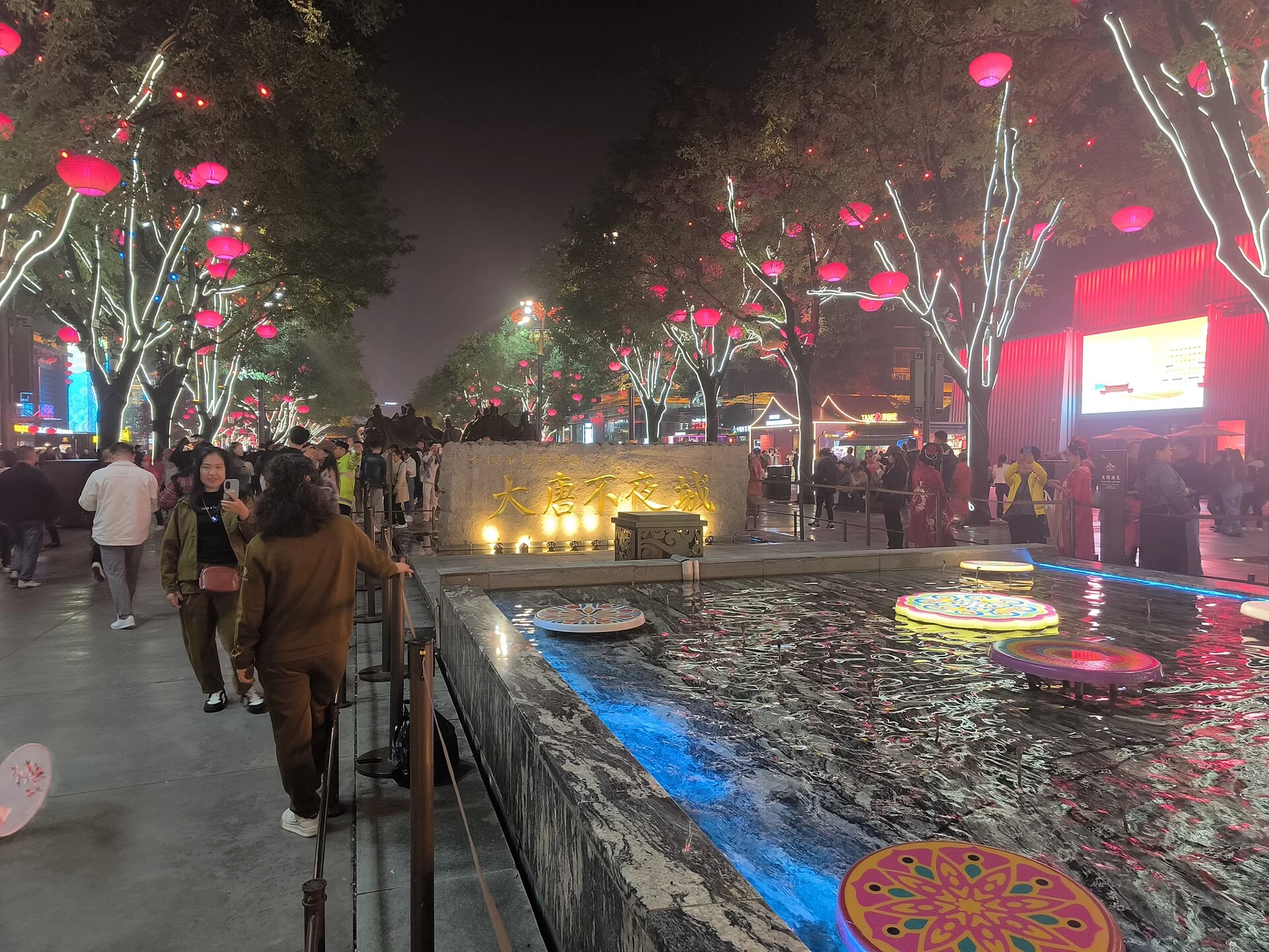 People walking beside a lit stone bearing the street name under red lanterns.