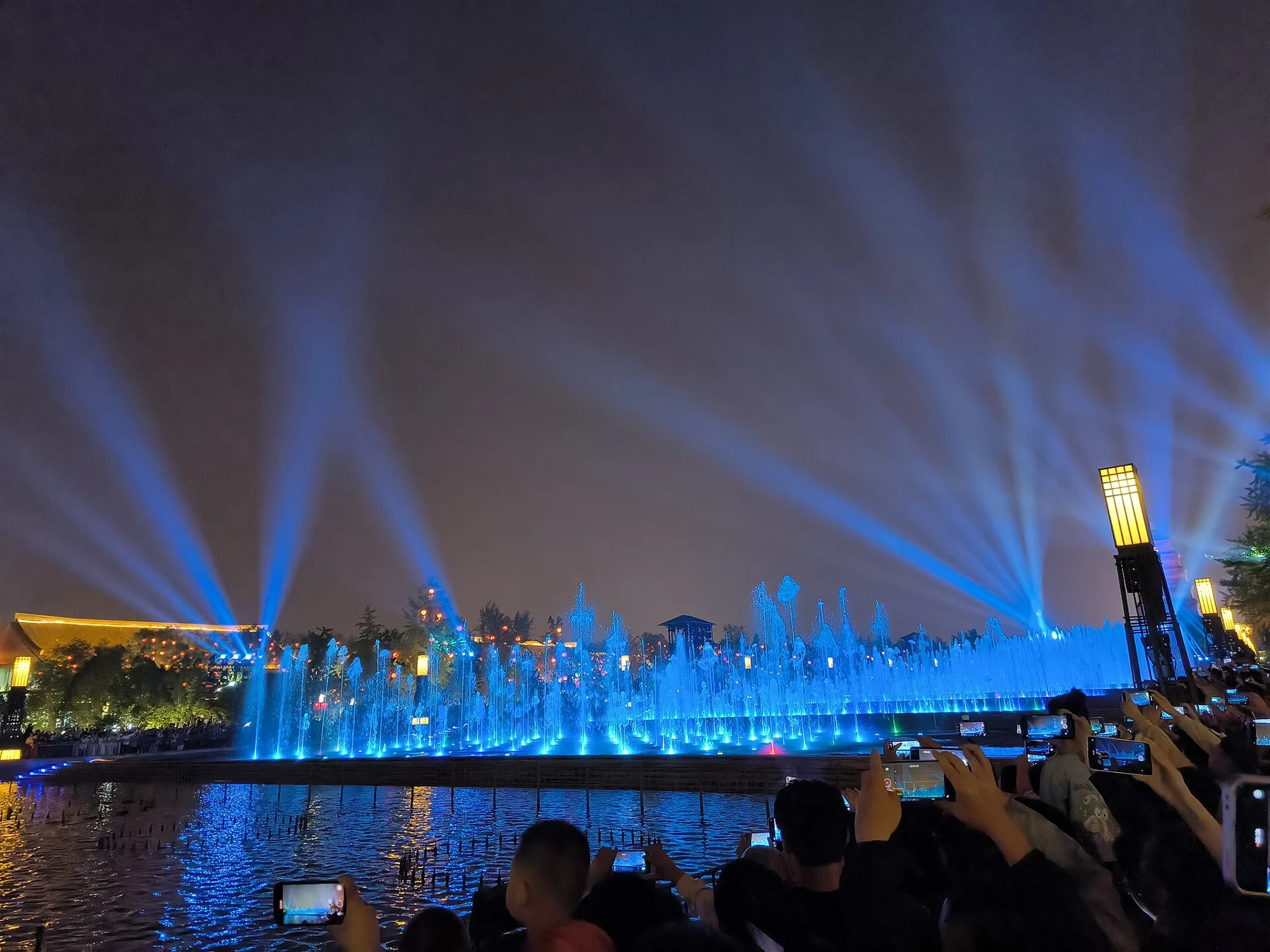 Blue-lit fountains and diagonal beams of light beyond a viewing crowd.